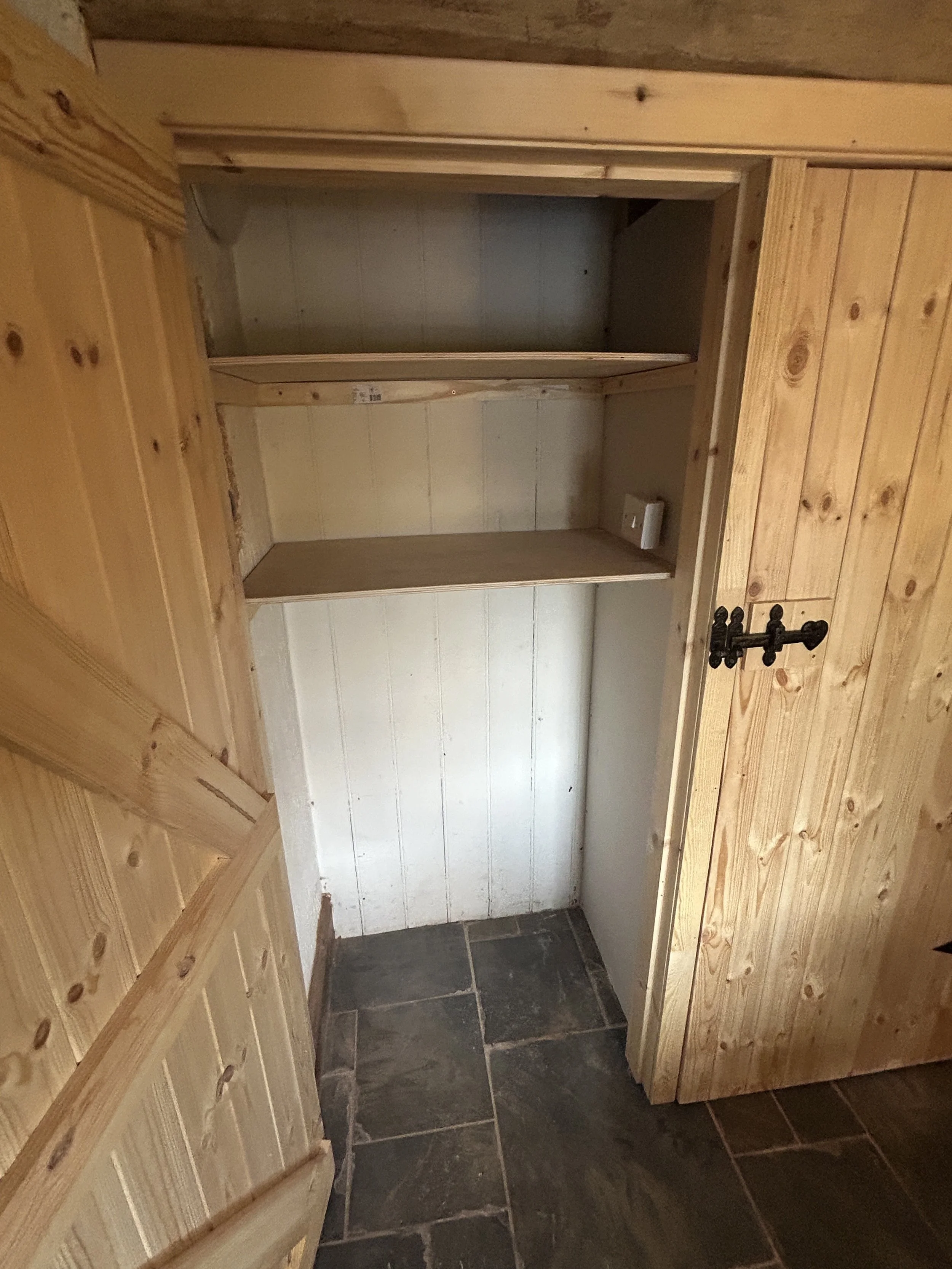 Interior view of a small, unfinished wooden closet or cabinet with a brick wall visible at the top, a white wall at the back, and dark floor tiles.