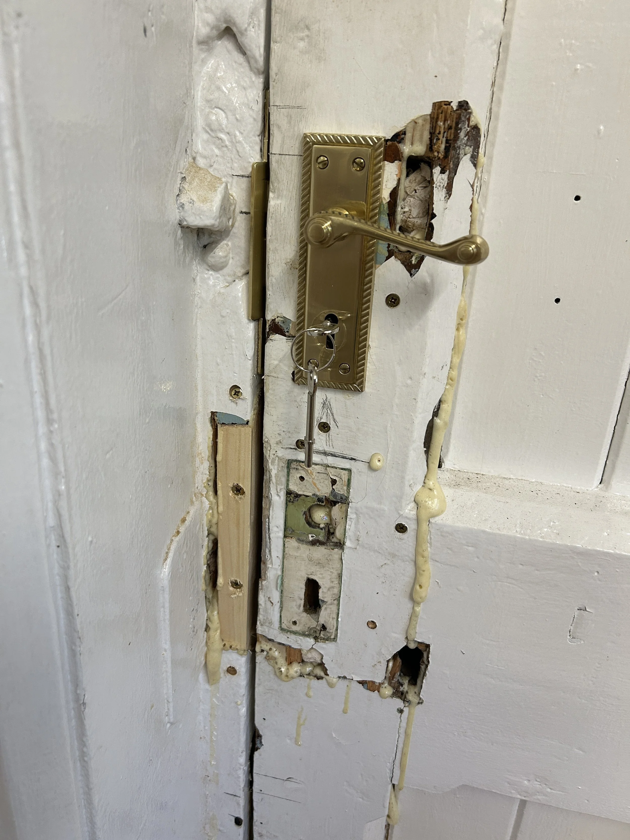 Close-up of a damaged door with an old, broken lock and exposed wood and wiring, showing deterioration and neglect.