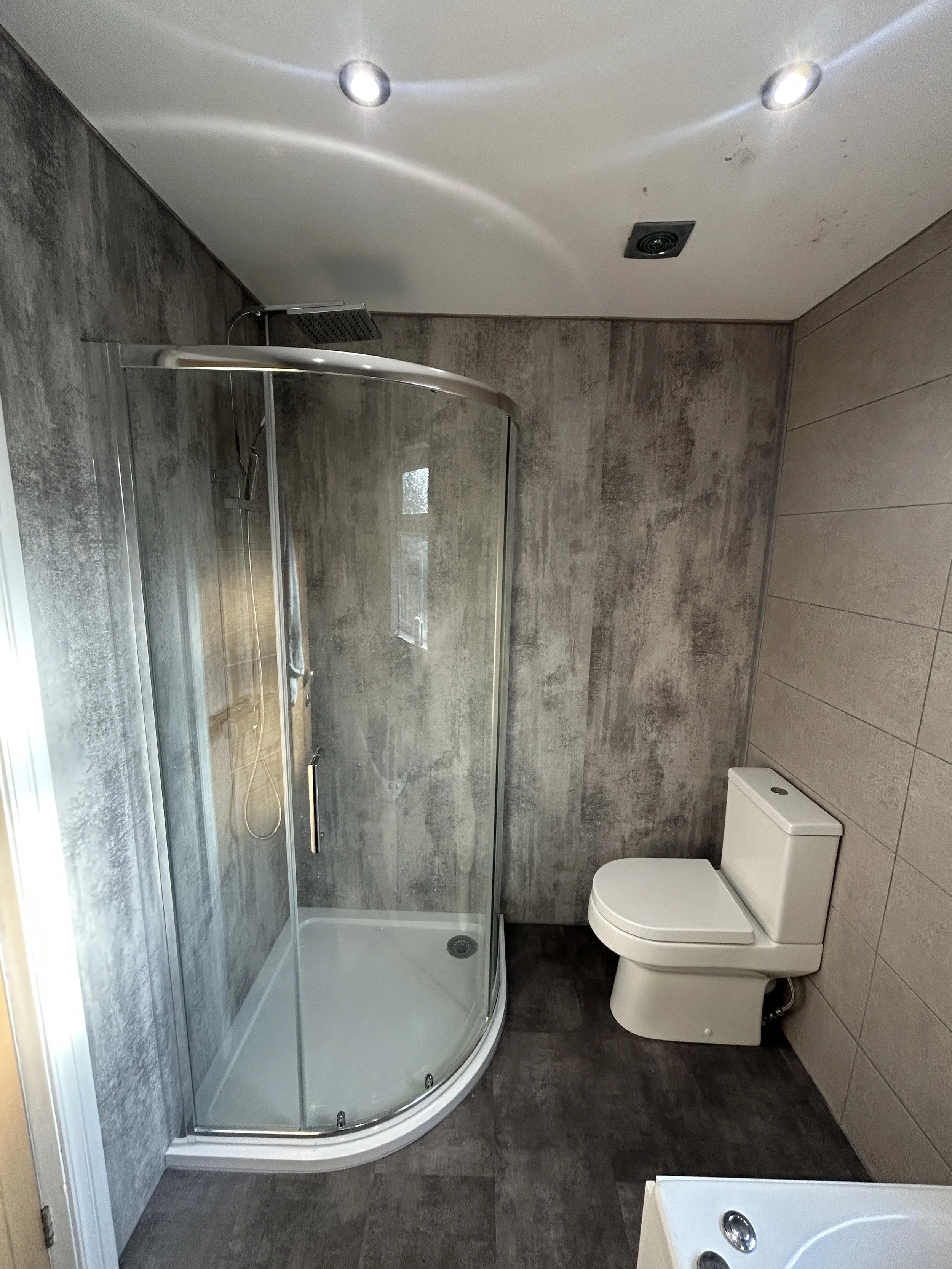Modern bathroom featuring a curved glass shower enclosure with a rainfall showerhead, a white toilet, a sink with two faucets, and gray textured wall tiles.