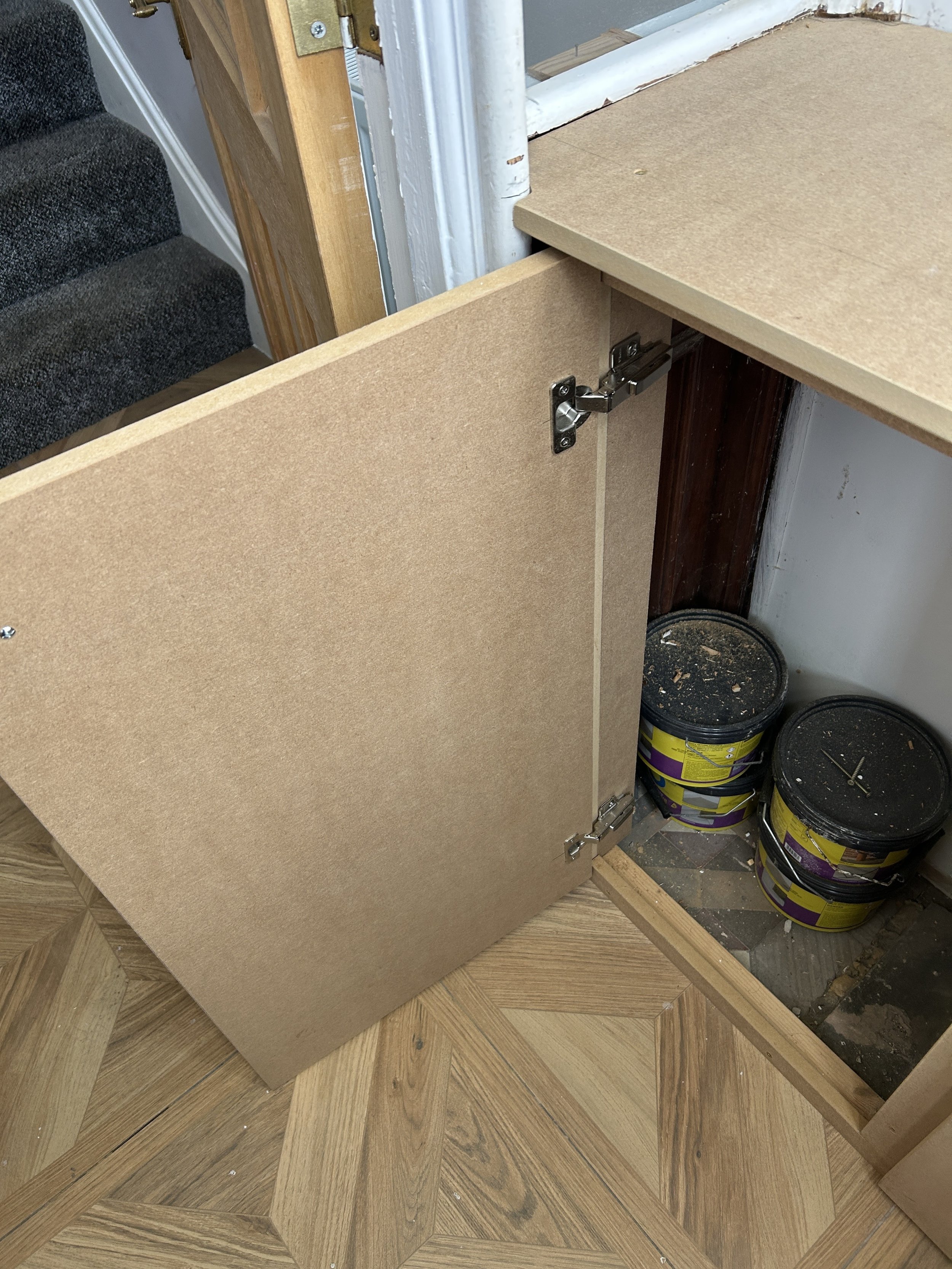 Partial view of a cabinet being assembled, showing the inside with open door and hardware, with paint buckets and a hardwood floor.