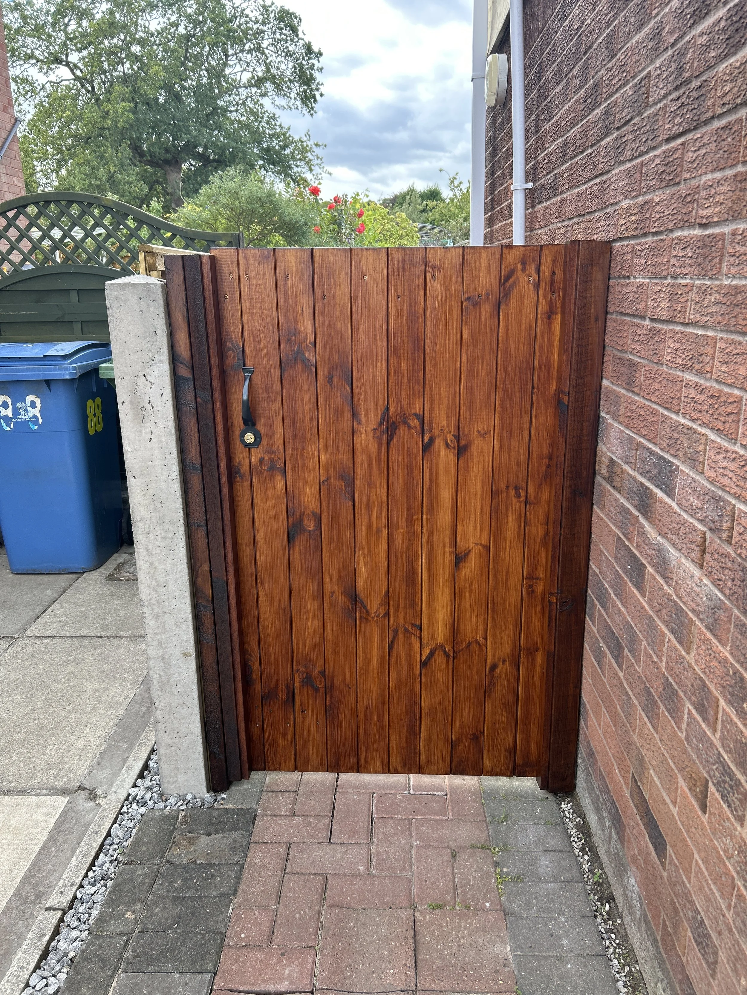 A wooden gate with a black handle, installed between a brick wall and a concrete post, with a brick paved ground in front and a garden with trees and flowers in the background.