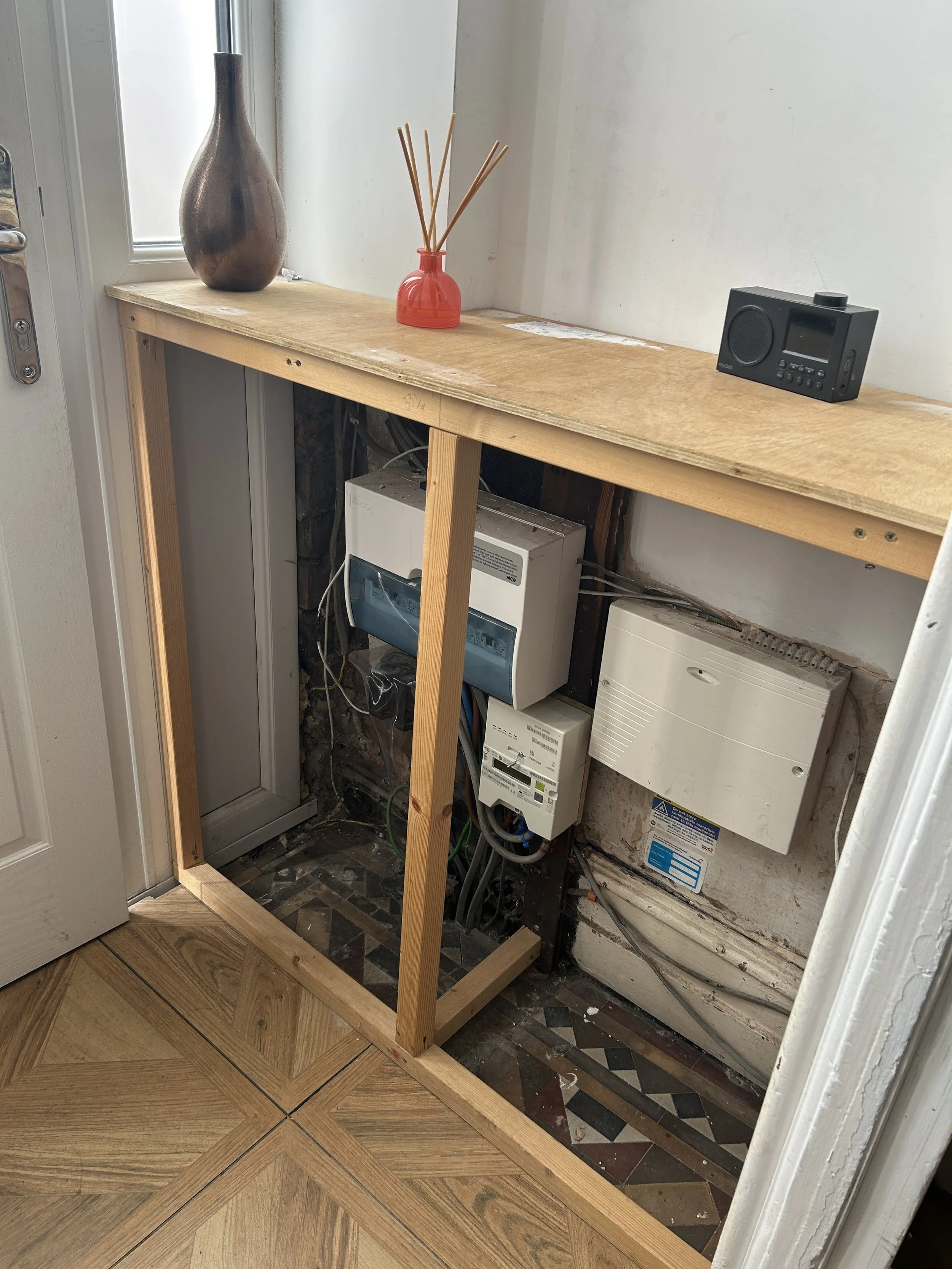 A partially constructed wooden frame inside a small utility space, with visible wiring, electrical boxes, and a small digital radio on a wooden shelf near a window, with a tiled floor and an unfinished wall.