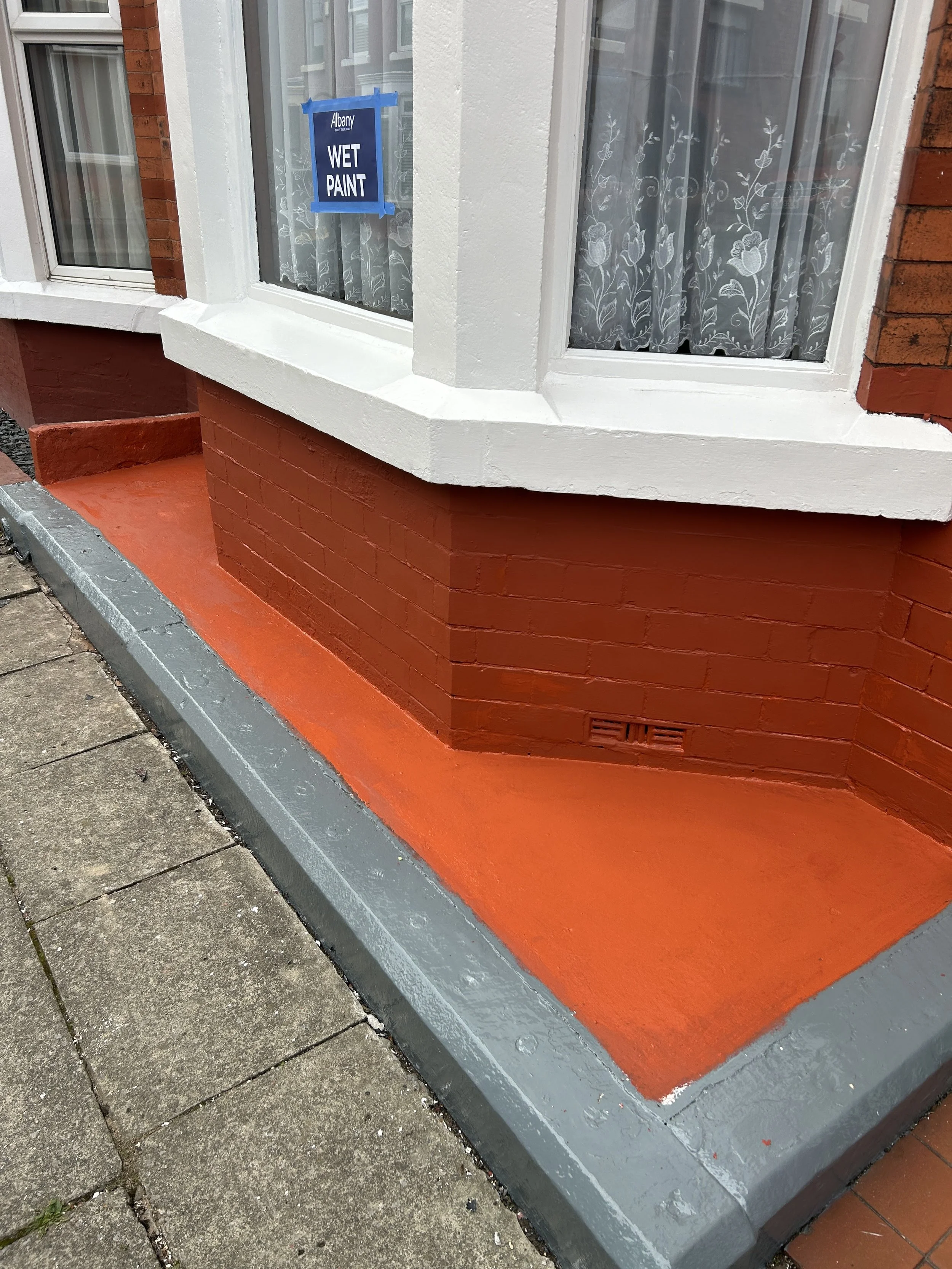 Close-up view of a house window with white framing, lace curtains with floral pattern, and a sign reading 'Wet Paint' taped on the window, with red brick wall and painted orange sidewalk outside.