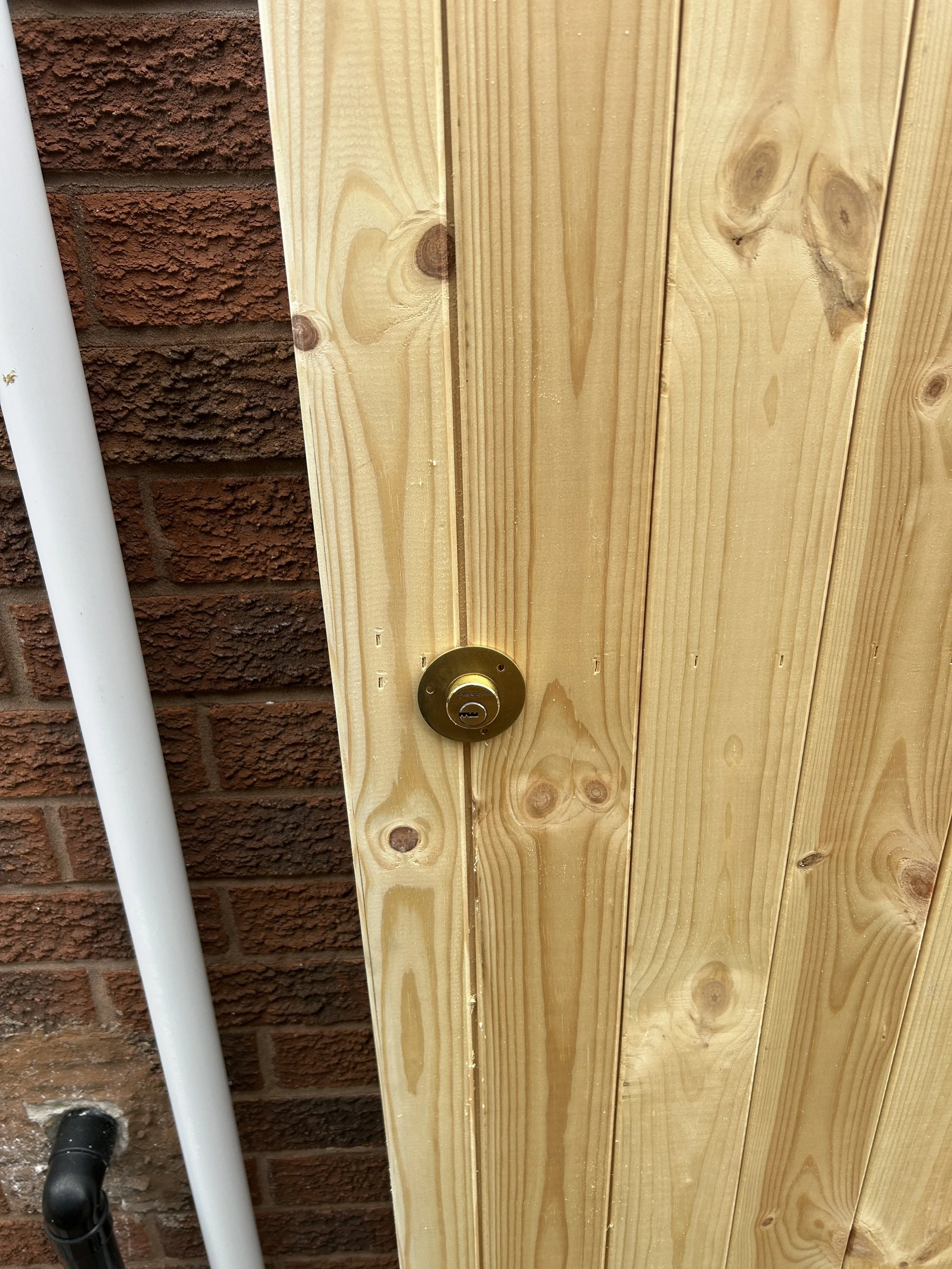 A wooden fence with a brass lock installed on it, and a brick wall with a white pipe adjacent to the fence.