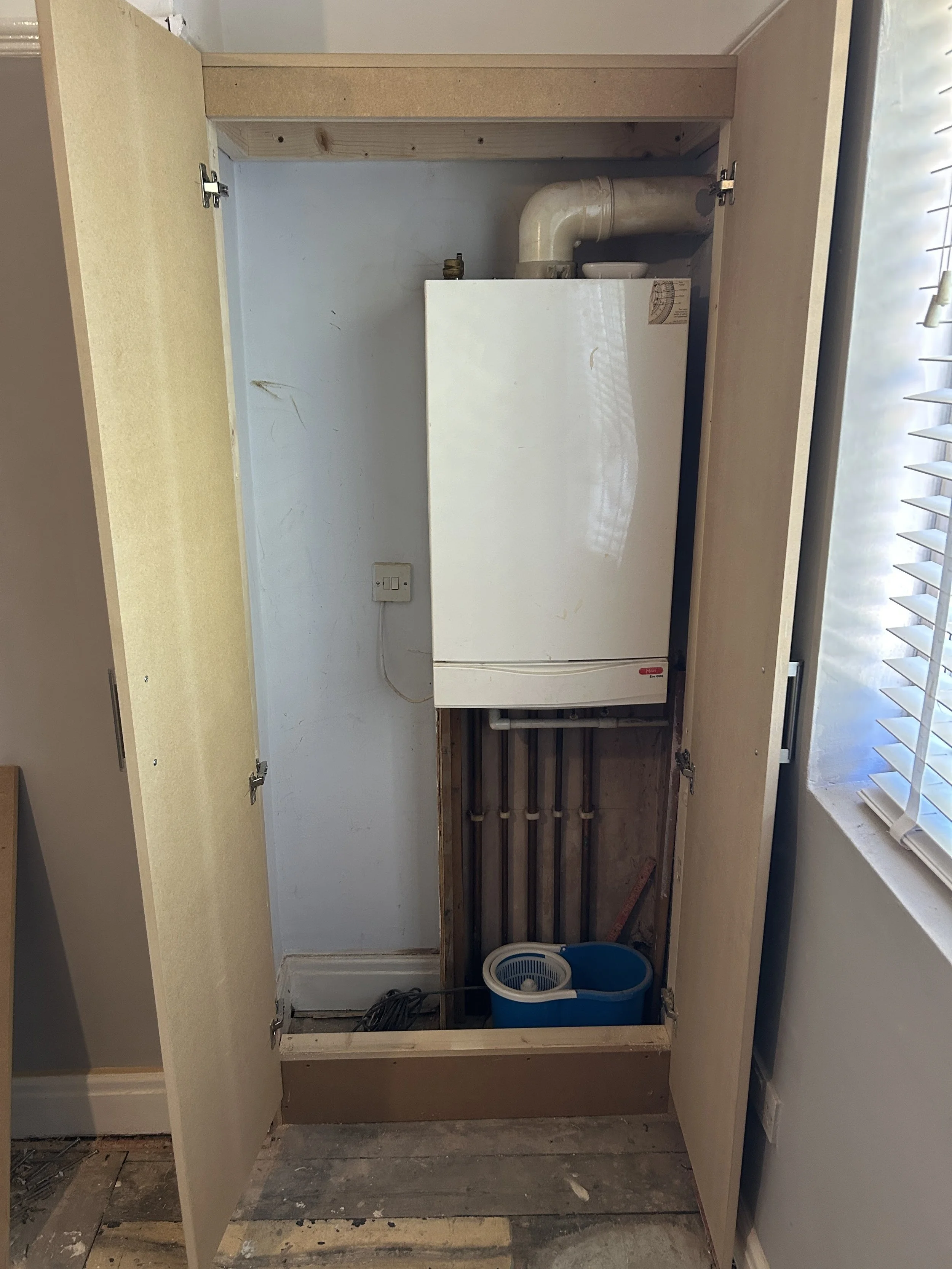 Kitchen cabinet with open doors, uncovering pipes and a small white appliance, possibly a water heater, with a blue trash can below and a window with blinds to the right.