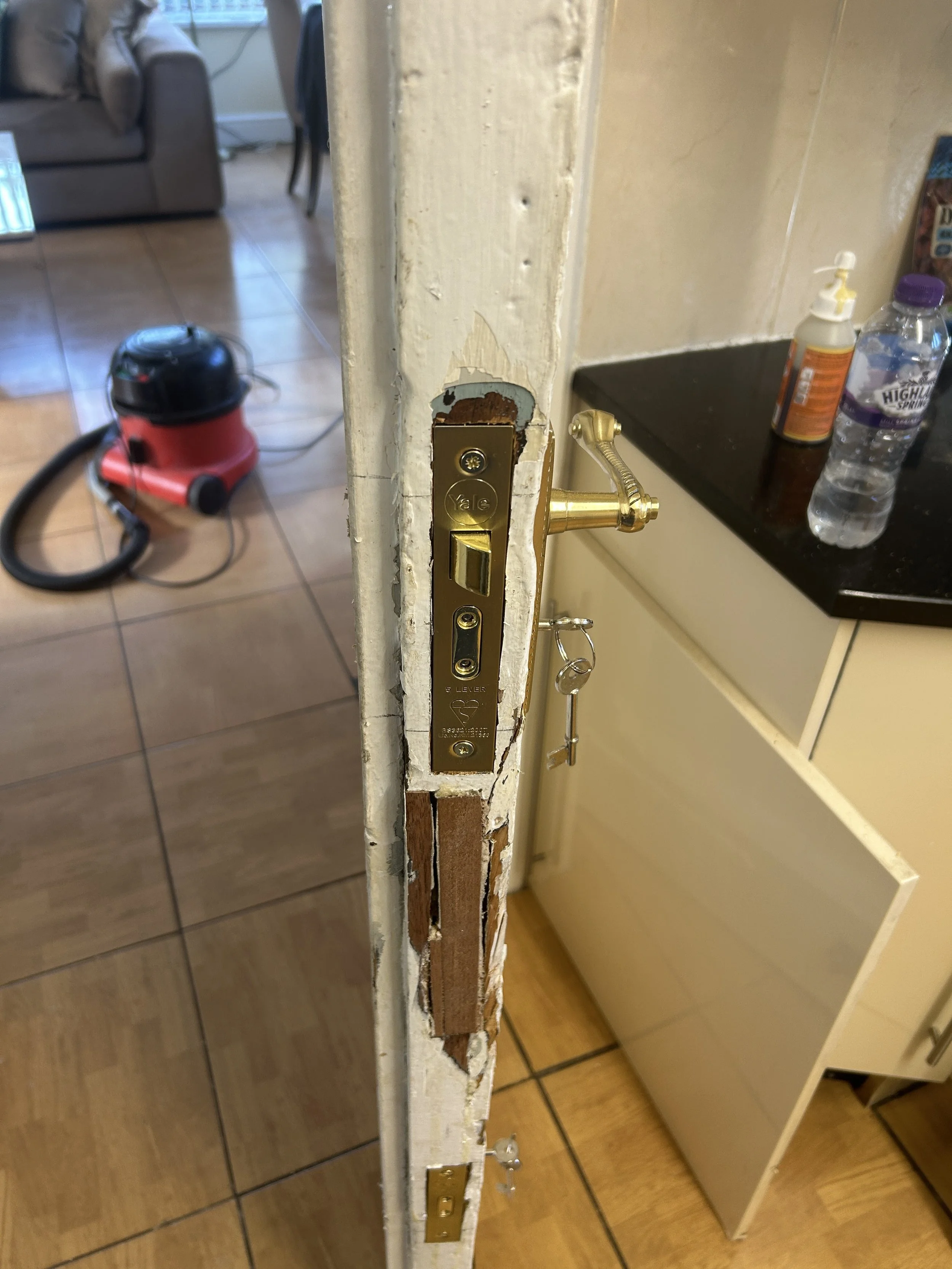 Close-up of a door latch and lockset installed on a door frame in a kitchen or dining area. The door frame is worn and partially stripped, revealing the wood beneath the paint.
