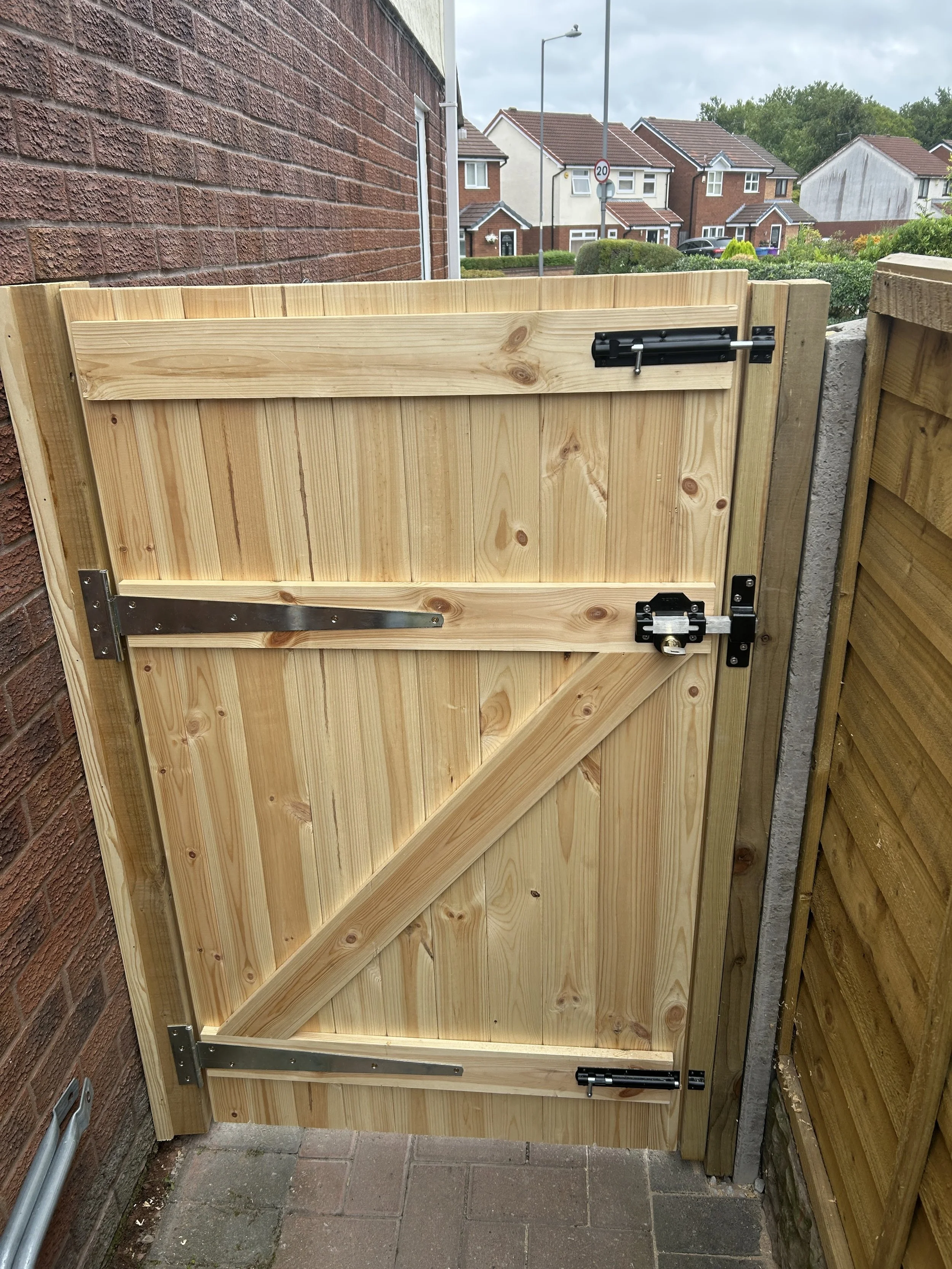 A wooden gate with metal hinges and a latch, installed between a brick wall and a wooden fence, on a paved pathway outside residential houses.