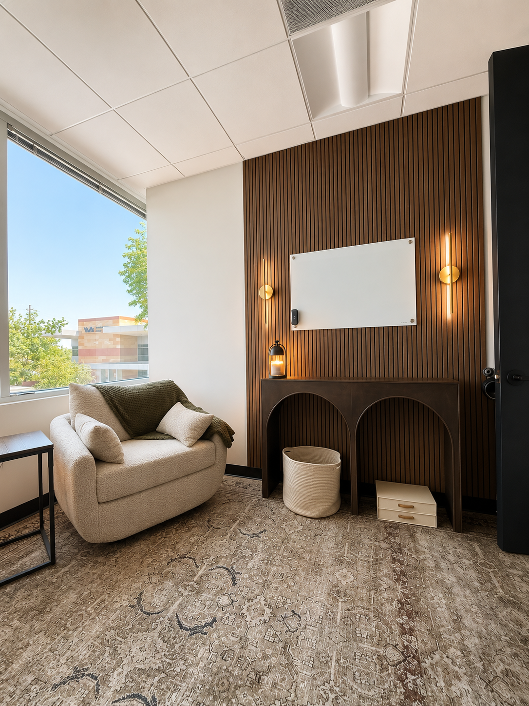 Cozy office corner with a beige armchair, green throw blanket, white pillows, small side table, wooden desk with arch-shaped openings, white basket, white drawer, whiteboard, wall-mounted lights, lantern, large window showing trees and buildings outside, patterned carpet, ceiling with tiles.