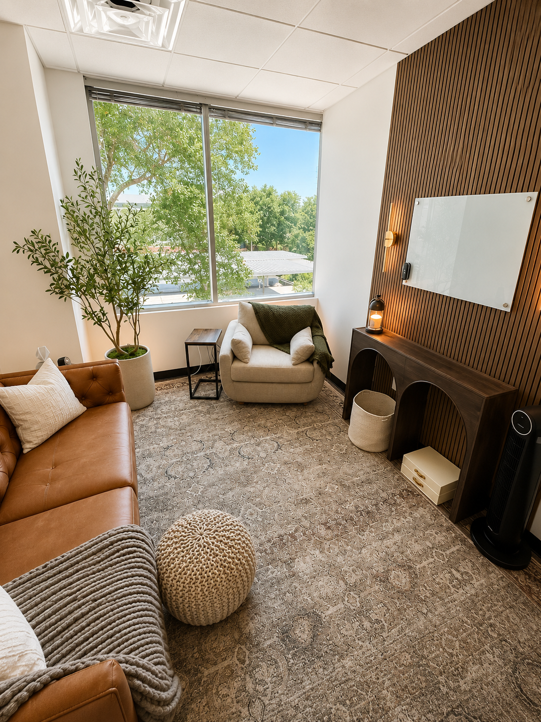 A cozy and modern office space with a large window showing greenery outside, decorated with a potted tree, a beige armchair with a green blanket, a brown leather sofa with cushions, a small wooden side table, a textured ottoman, a wooden console with a lamp, a whiteboard, and a fan, all on an intricate patterned rug.