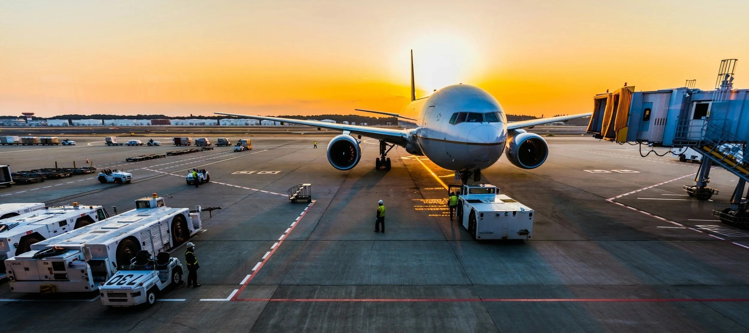 Passenger airplane parked at gate with jet bridge connected, airport vehicles and crew members on tarmac during sunset.
