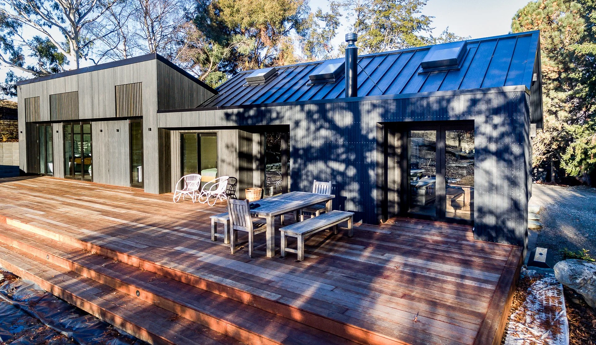 Modern dark-gray cabin with a blue metal roof, wood deck, outdoor dining table, lounge chairs, and surrounding trees