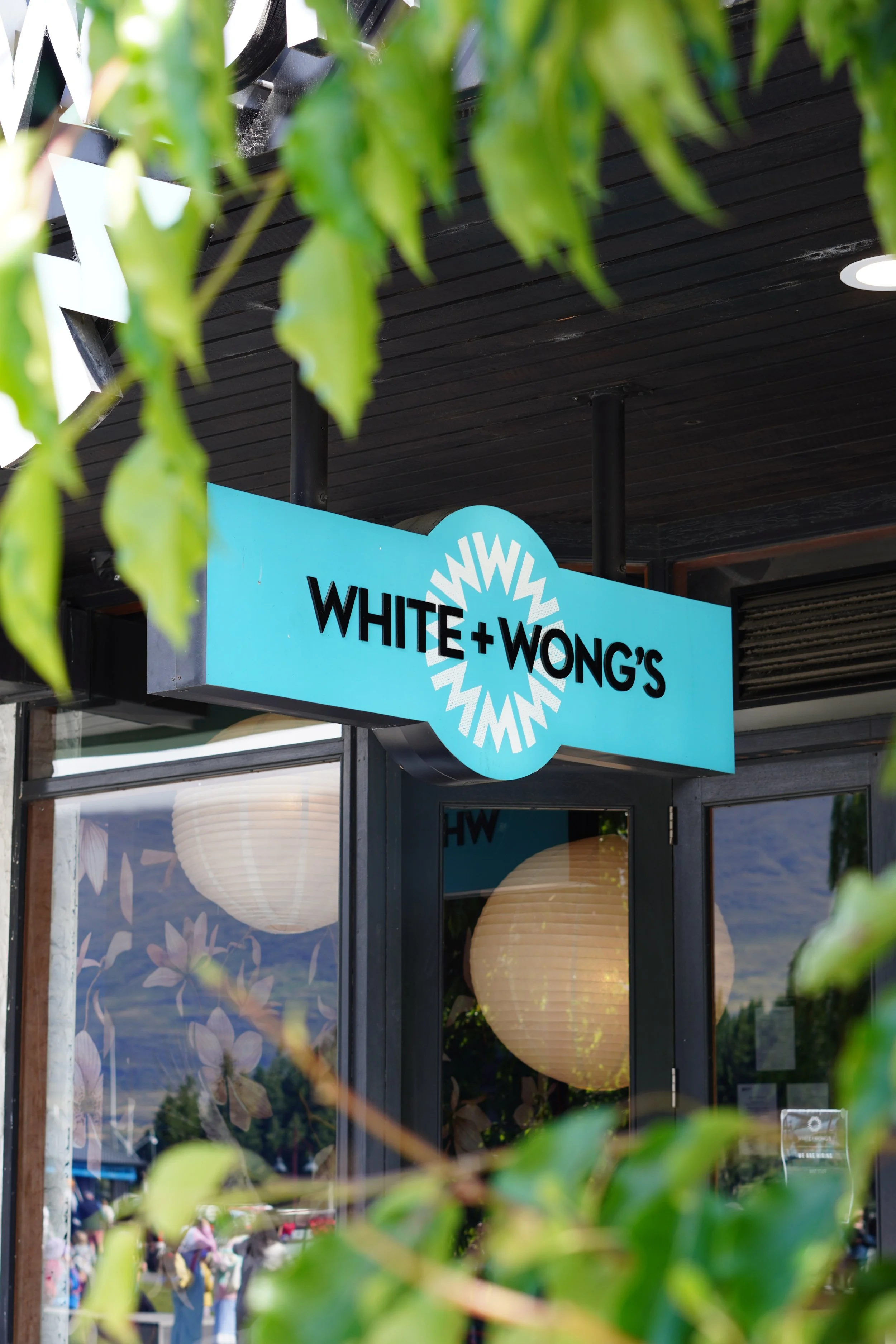 Turquoise White + Wong’s restaurant sign partially obscured by green leaves outside a black storefront.
