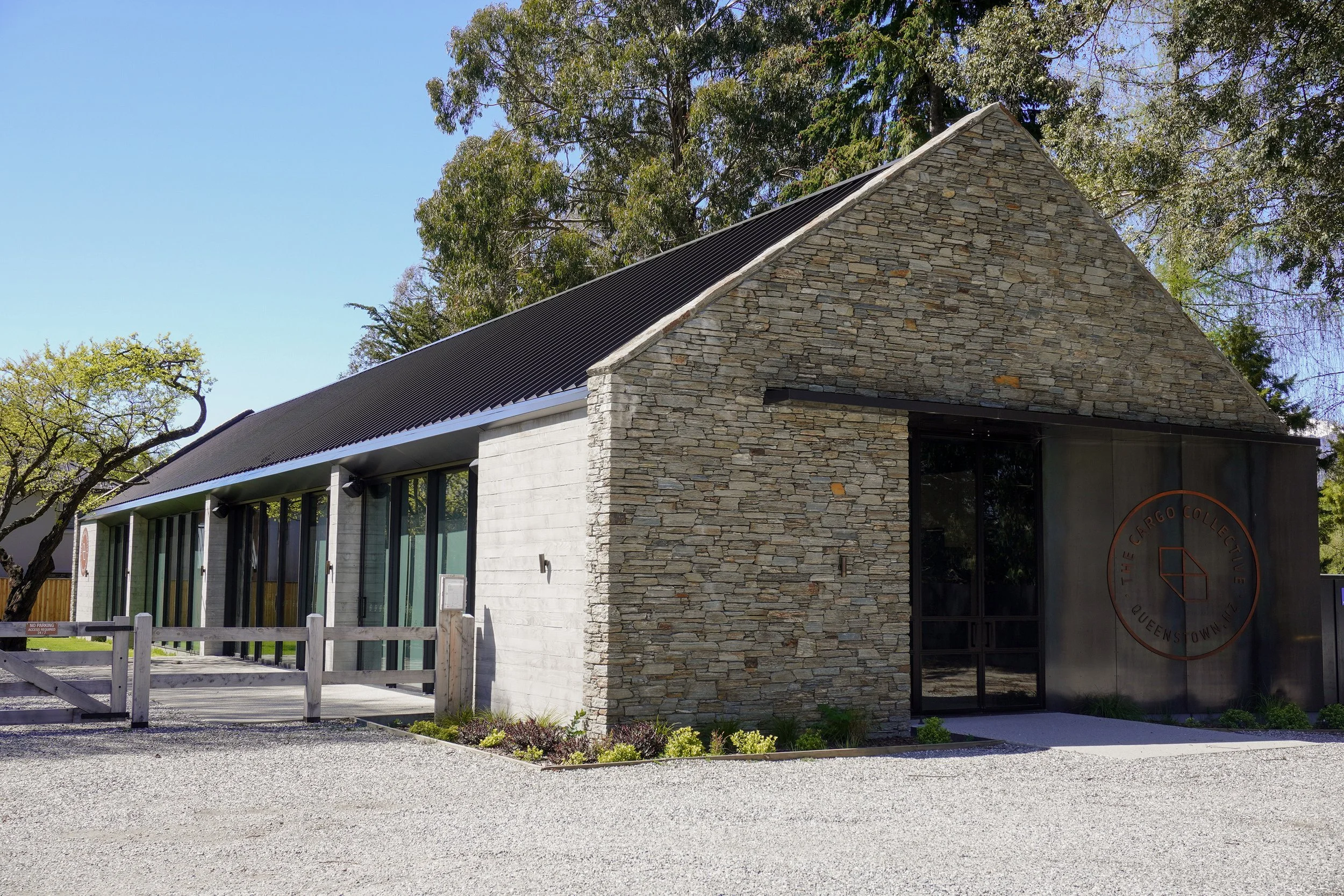 Exterior of The Cargo Collective building in Queenstown, New Zealand, featuring a stone facade, black metal roof, trees, and landscaped grounds.