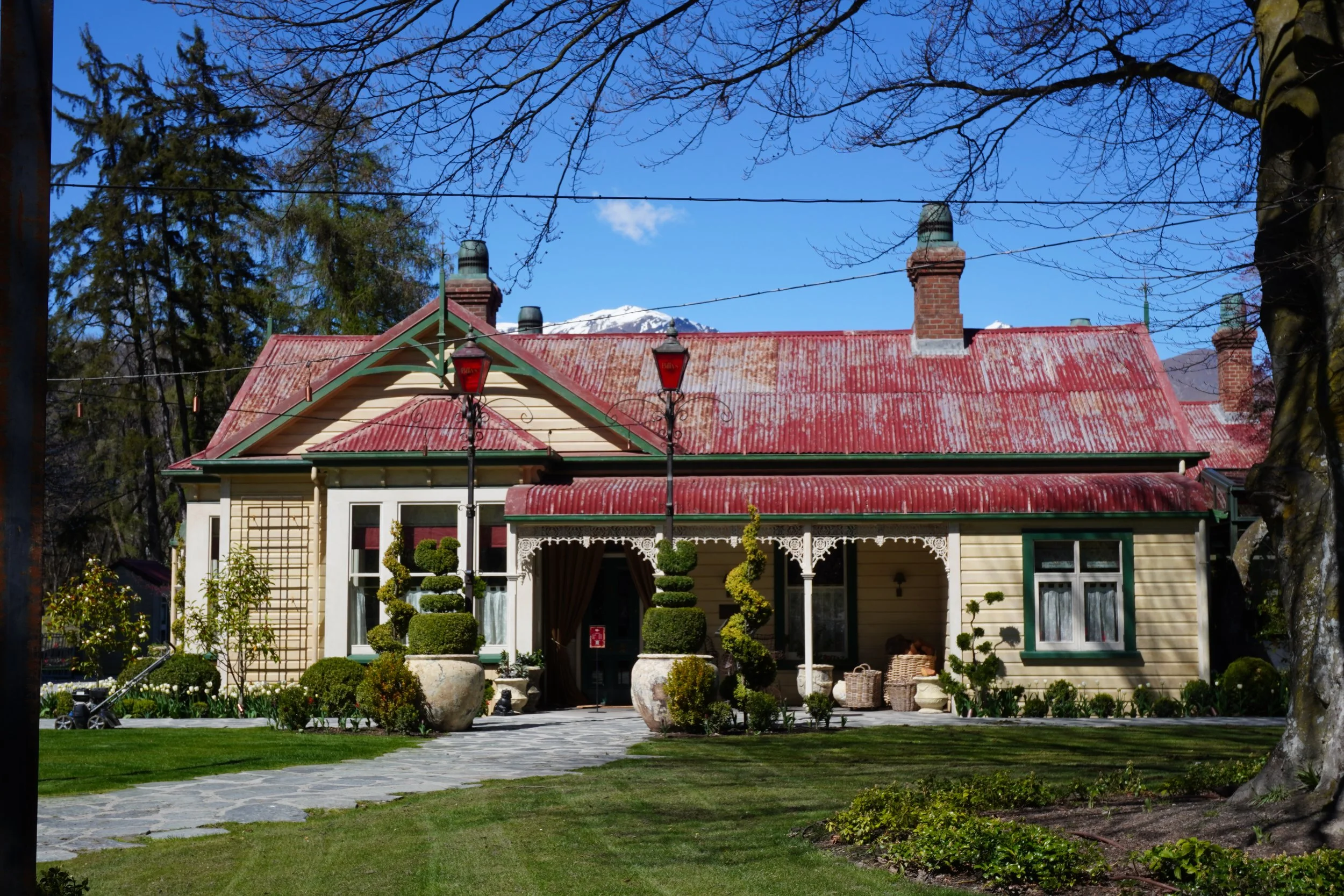 Historic Victorian-style building with ornate verandah, manicured gardens, red corrugated roof, and snow-capped mountains in the background
