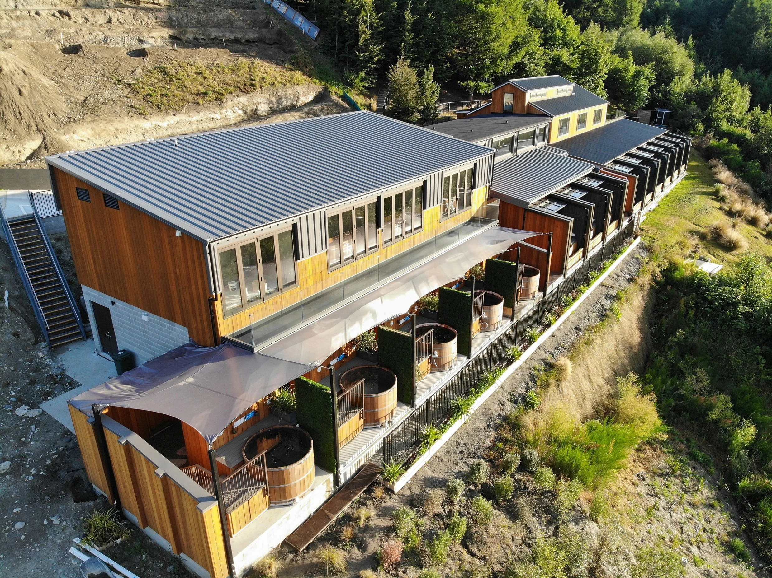 Aerial view of a modern multi-unit lodge with metal roofs, wooden siding, private balconies, and outdoor soaking tubs nestled in a forested hillside.