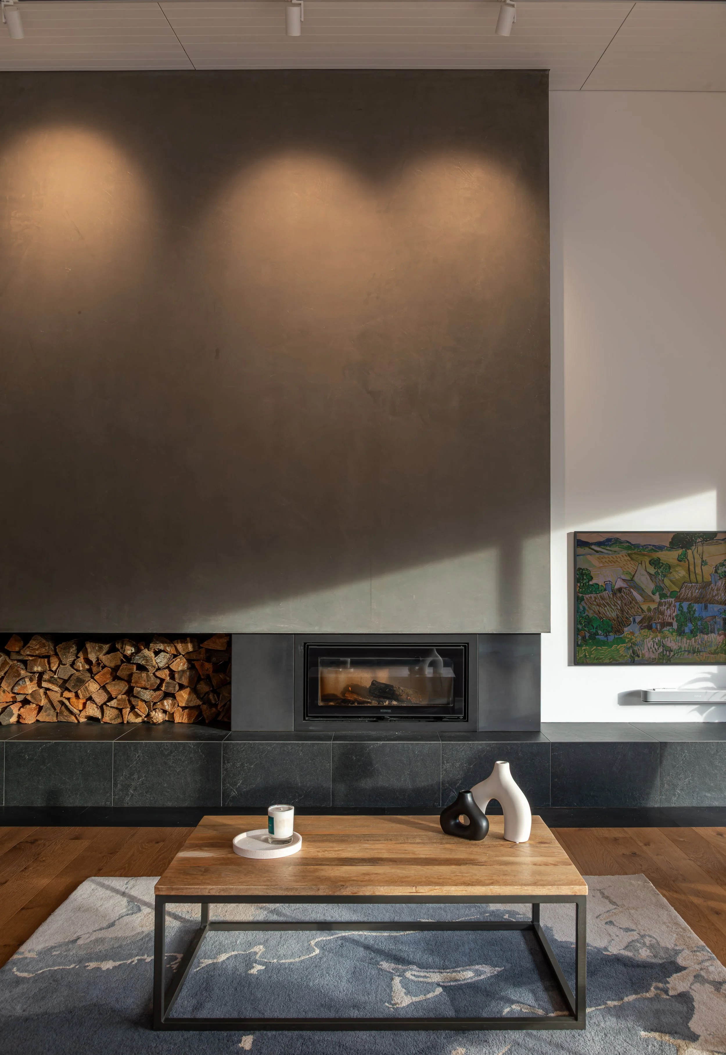 Modern living room with a large dark fireplace, stacked firewood, landscape painting, wooden coffee table, ceramic vases, and patterned area rug