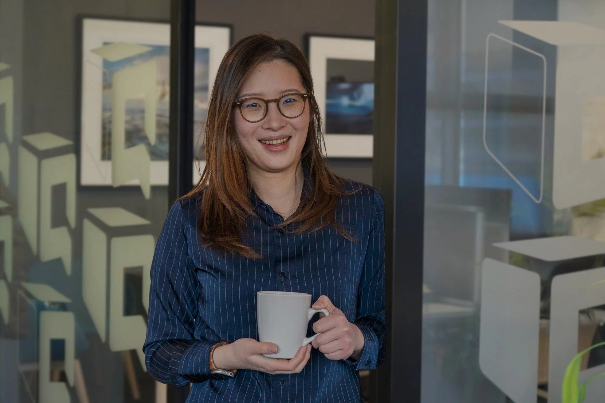 Smiling woman wearing glasses and a navy striped shirt while holding a white coffee cup in an office