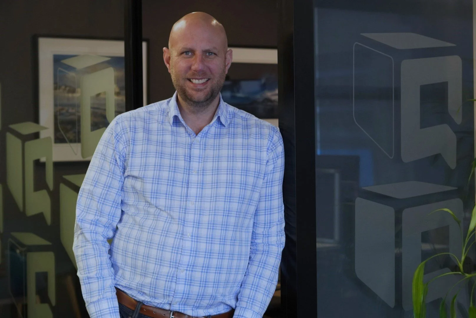Smiling bald man in a blue plaid shirt standing in a modern office beside framed artwork and a glass partition.