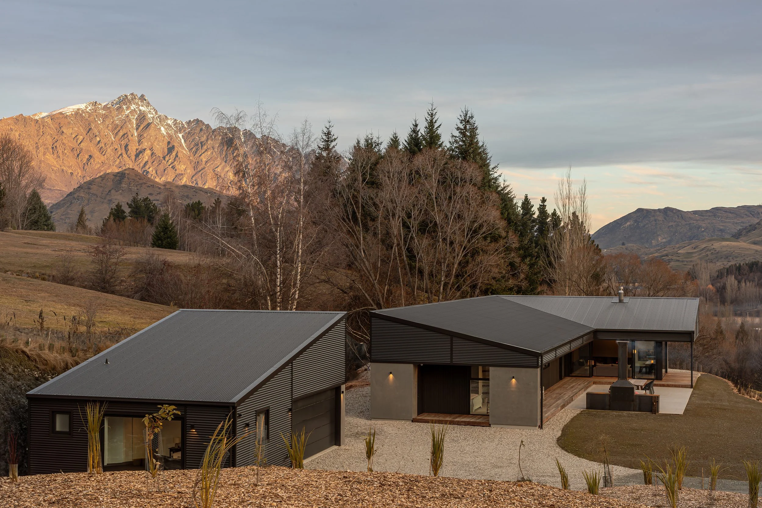 Modern black metal-clad house with corrugated roofs overlooking snow-capped mountains, evergreen trees, and a valley at sunset