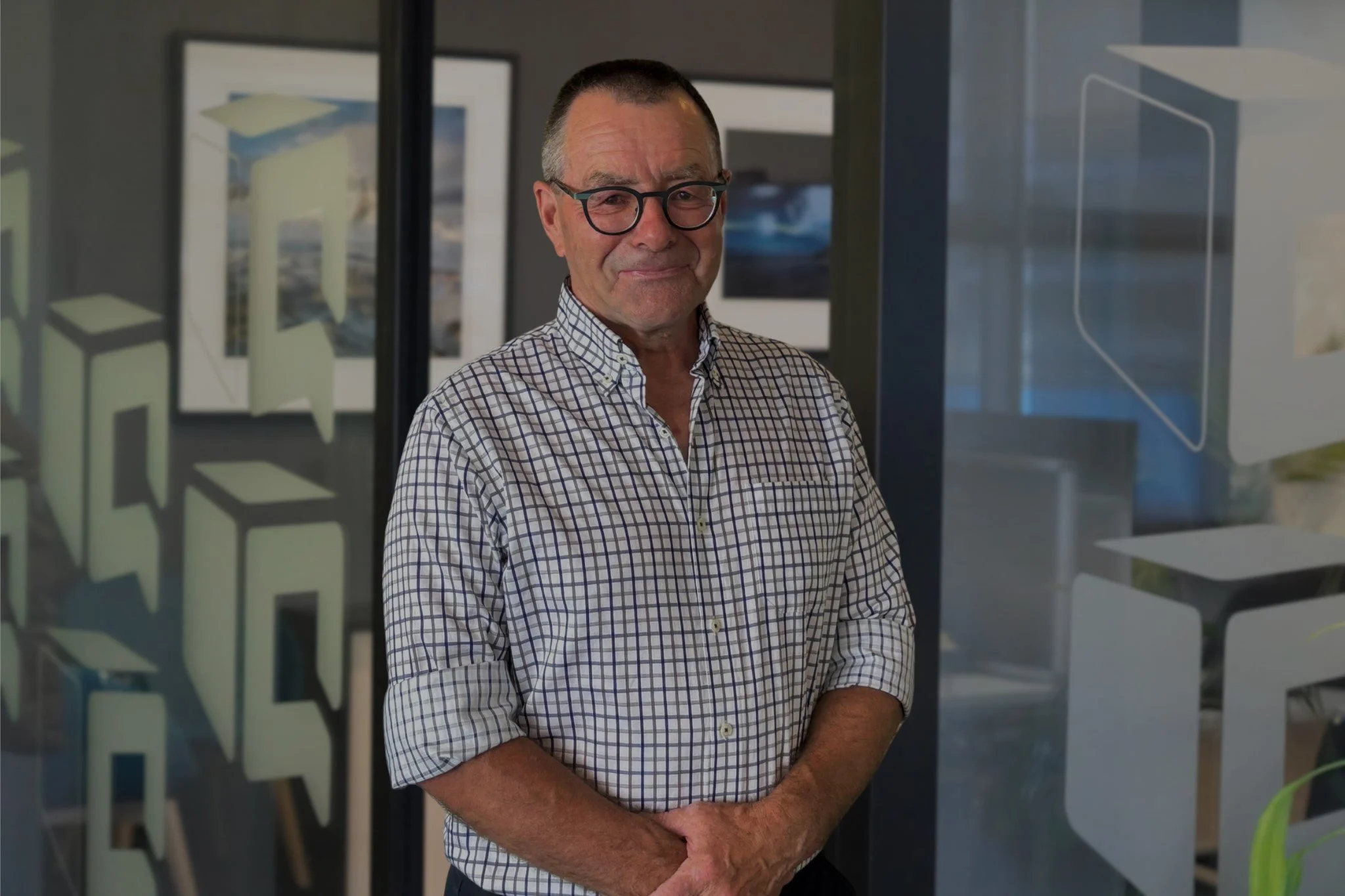 Smiling older man wearing glasses and a checkered shirt standing in a modern office with framed artwork.