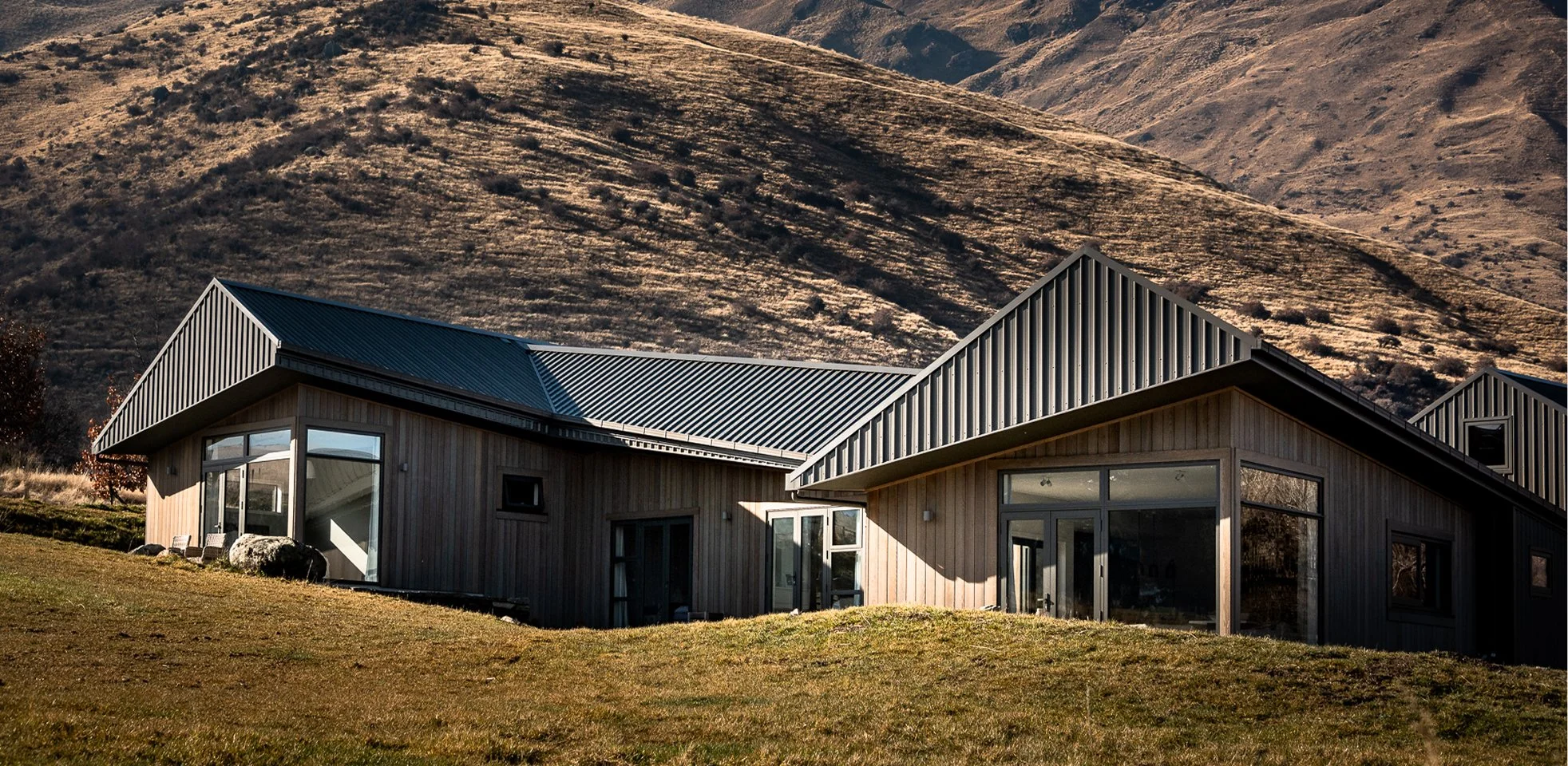 Modern wood-clad house with metal roofs on a grassy hillside surrounded by dry mountain slopes