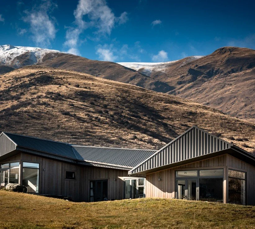 Modern lodge with metal roofs in front of snow-dusted brown mountains under a blue sky