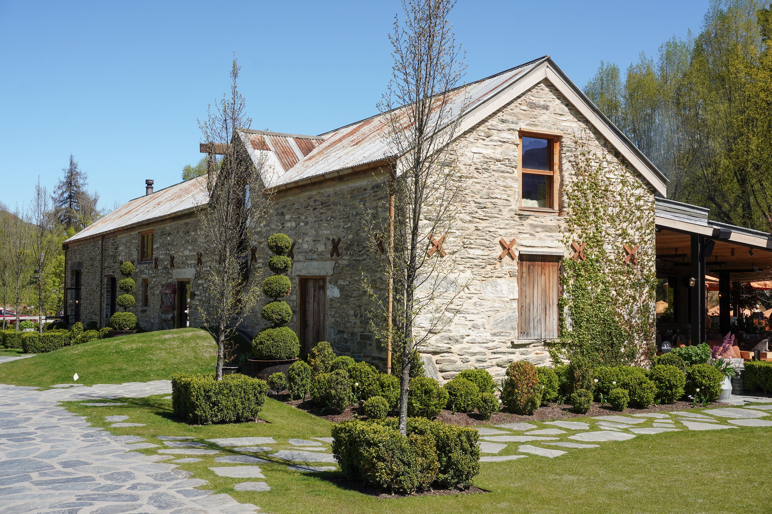 Rustic stone building with a weathered metal roof, manicured boxwood gardens, and slate walkways beneath a clear blue sky