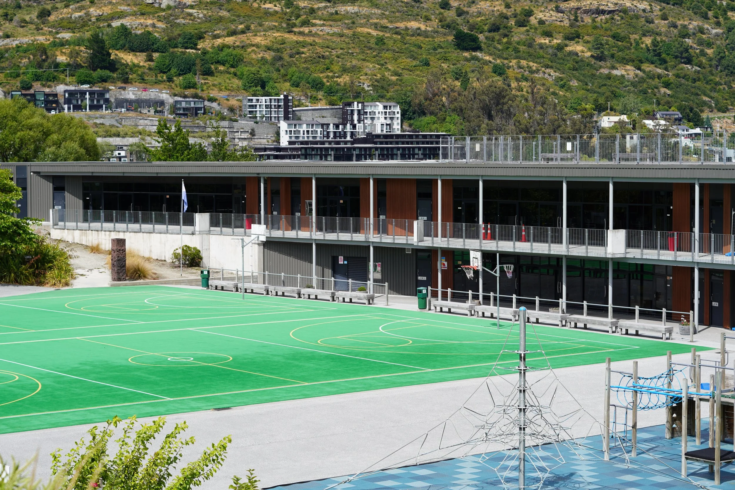 Modern school campus with a green multi-sport court, basketball hoops, playground equipment, and hillside apartments in the background