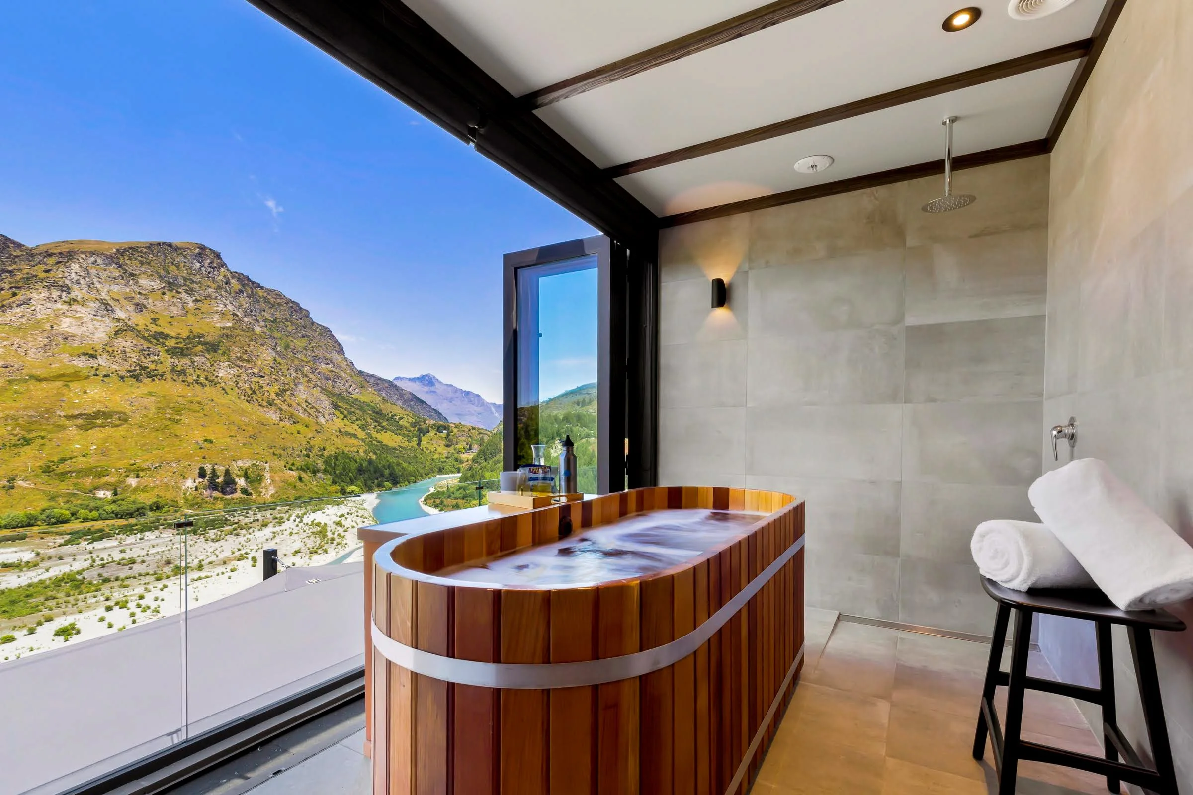 Luxury bathroom with a wooden soaking tub, rainfall shower, and panoramic mountain and turquoise lake views