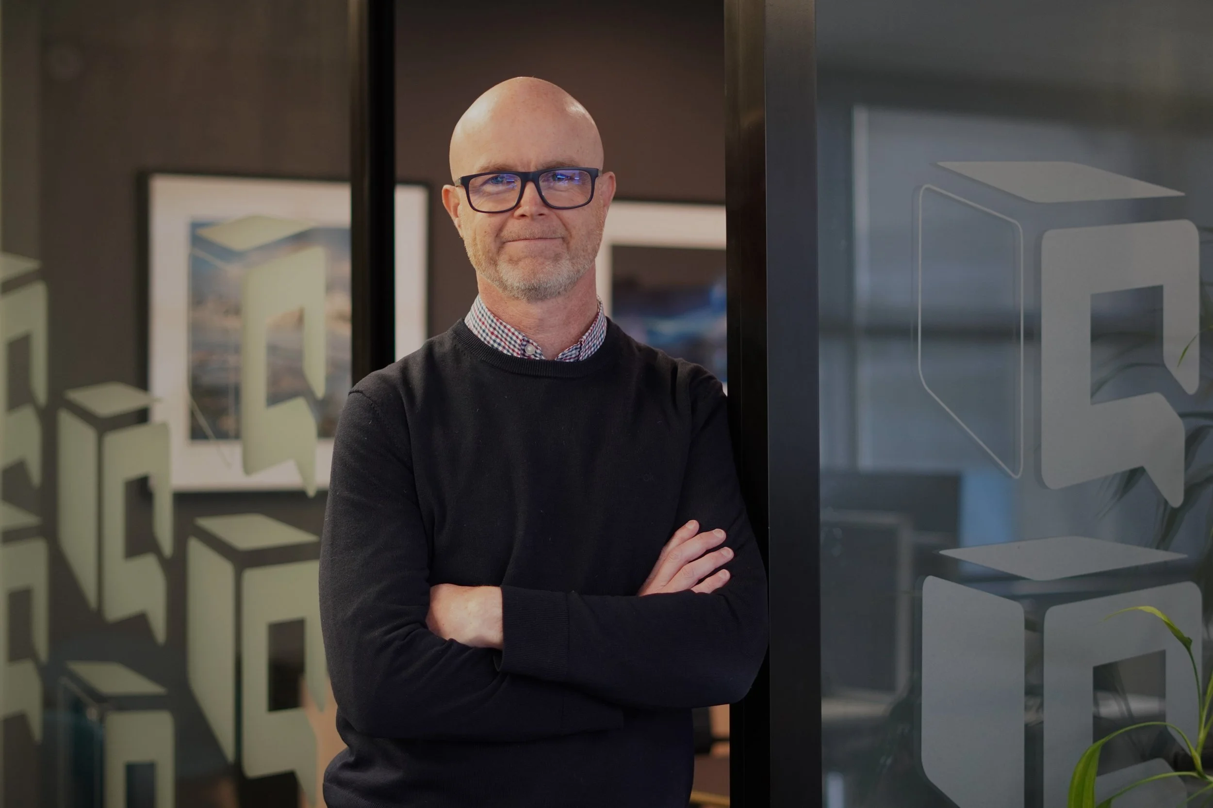 Bald man wearing glasses and a black sweater stands with folded arms in a modern office.