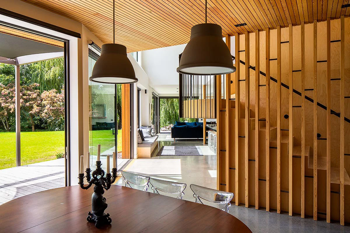 Modern open-plan dining room with wood-paneled walls, pendant lights, glass chairs, and garden views