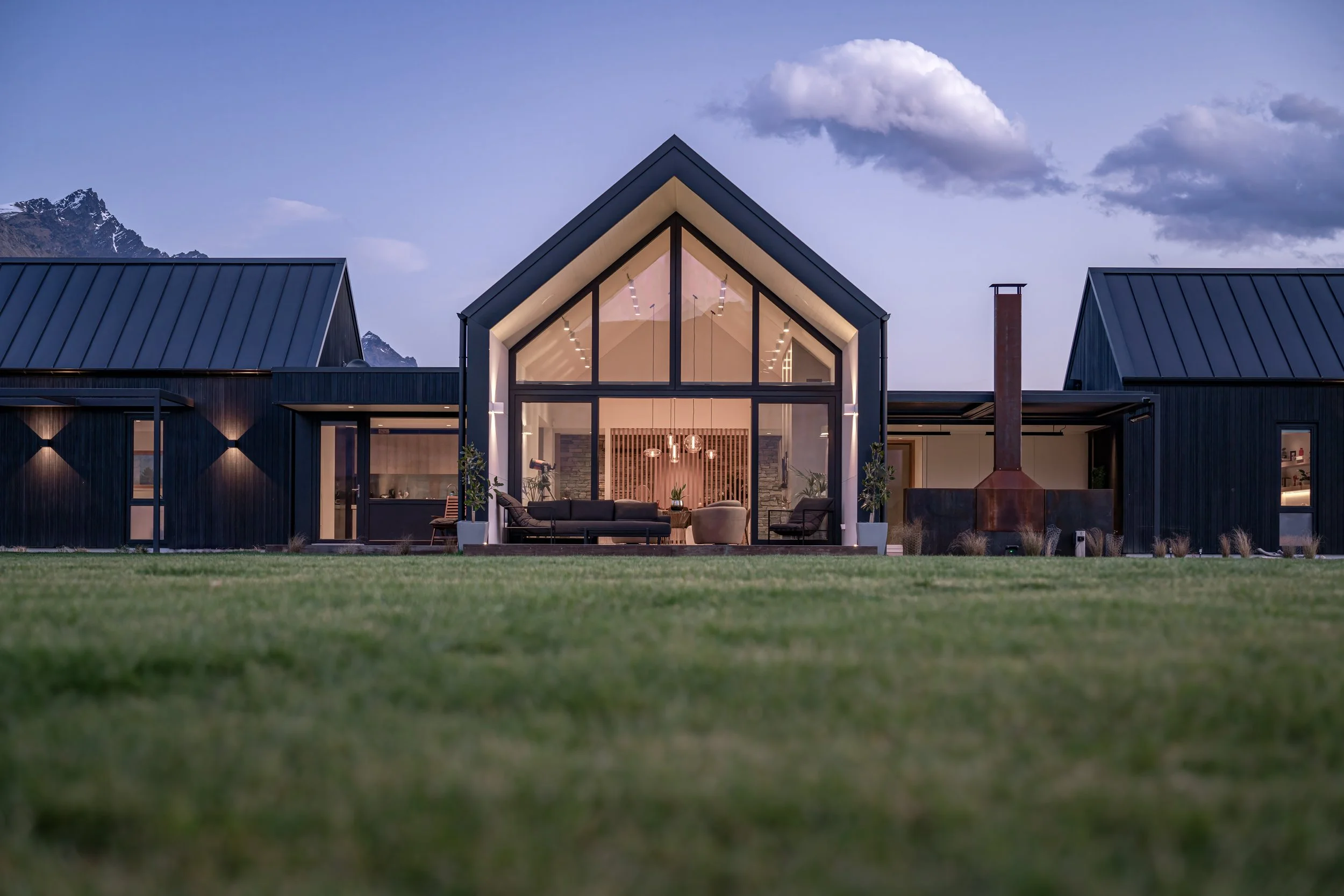Modern black metal mountain home with floor-to-ceiling windows, outdoor fireplace, and illuminated interior at dusk