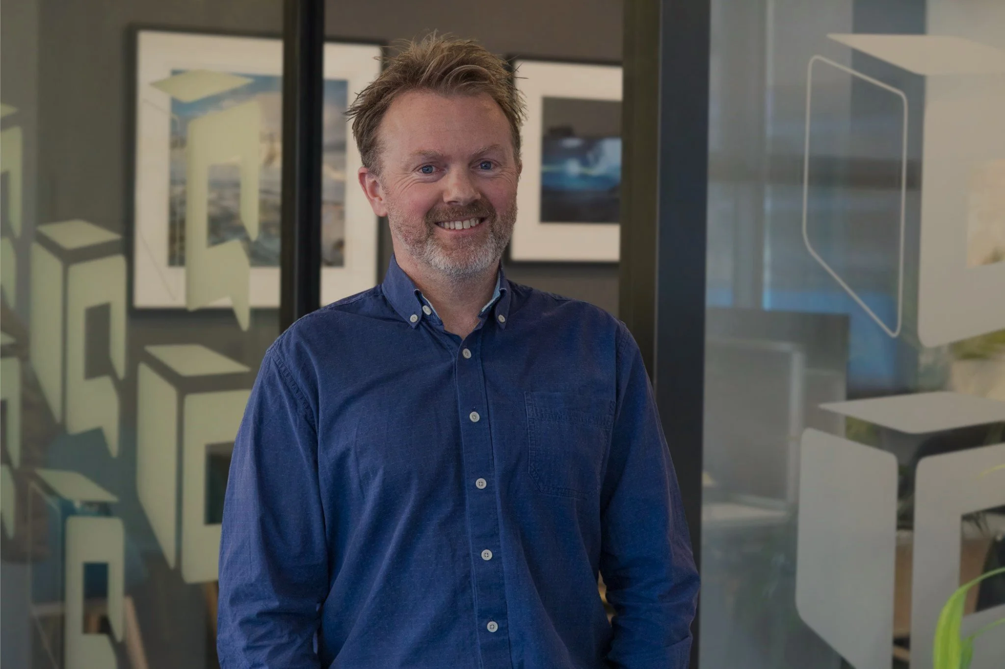 Smiling man in a blue button-down shirt standing in a modern office with framed artwork.