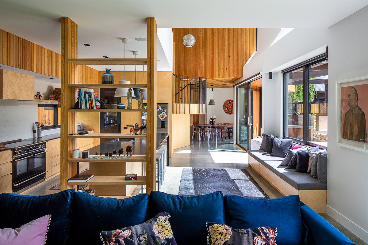 Modern open-plan home interior with wood-paneled walls, kitchen, shelving divider, spiral staircase, dining area, and blue sectional sofa