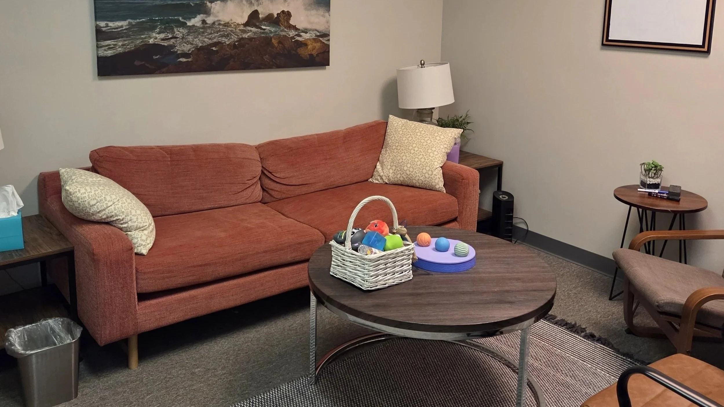Living room with a burnt orange sofa, a round wooden coffee table with a basket of colorful toys, a side table with a lamp, and wall art depicting a coastline. Small side table with plants and electronics, framed artwork on the wall, and a carpeted floor.