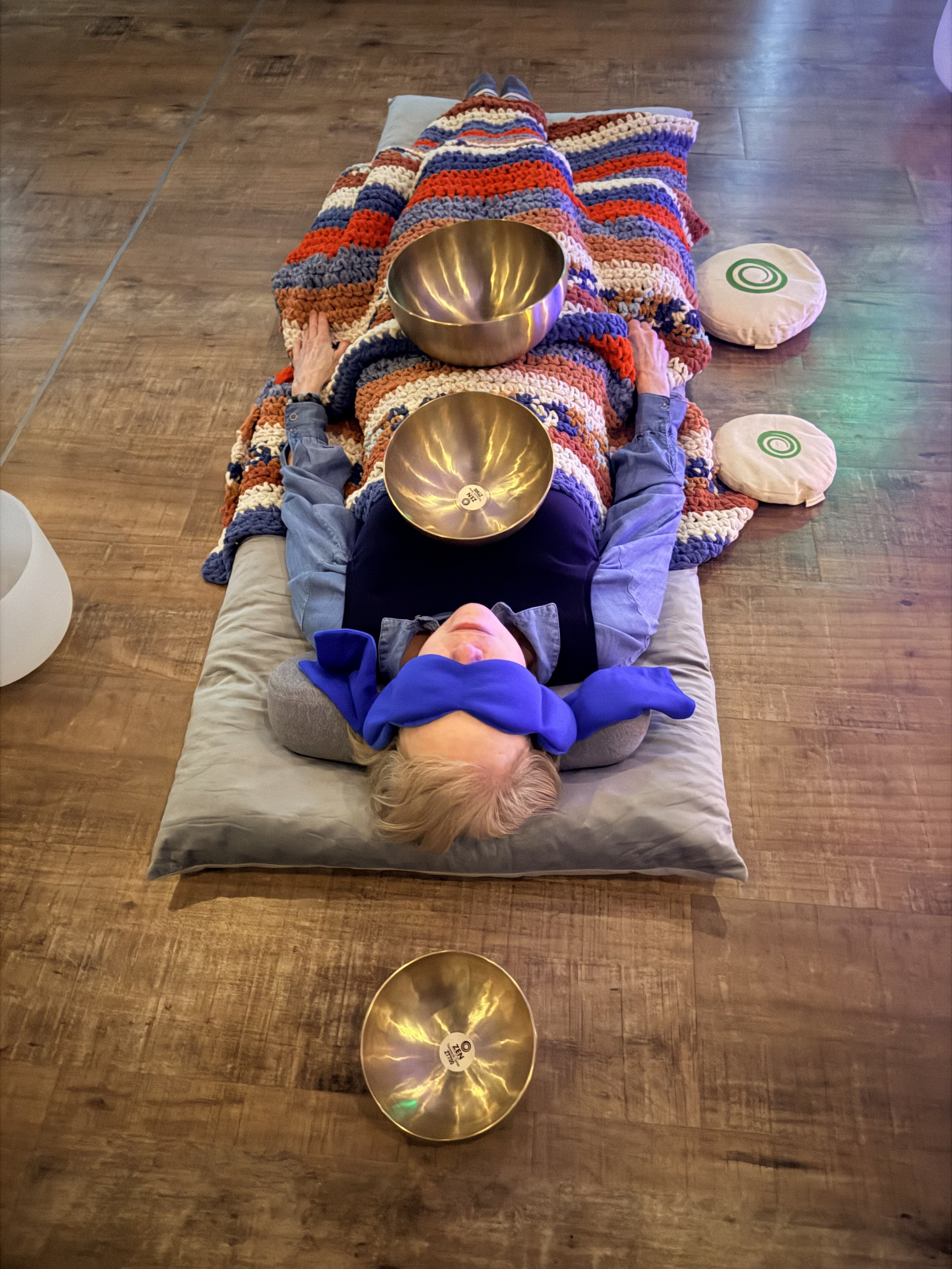 A person lying on their back on a floor with two singing bowls resting on their stomach, while wearing a multicolored crocheted blanket. Their head is covered with a blue cloth, and there are two additional bowls. Private meditation therapy transform