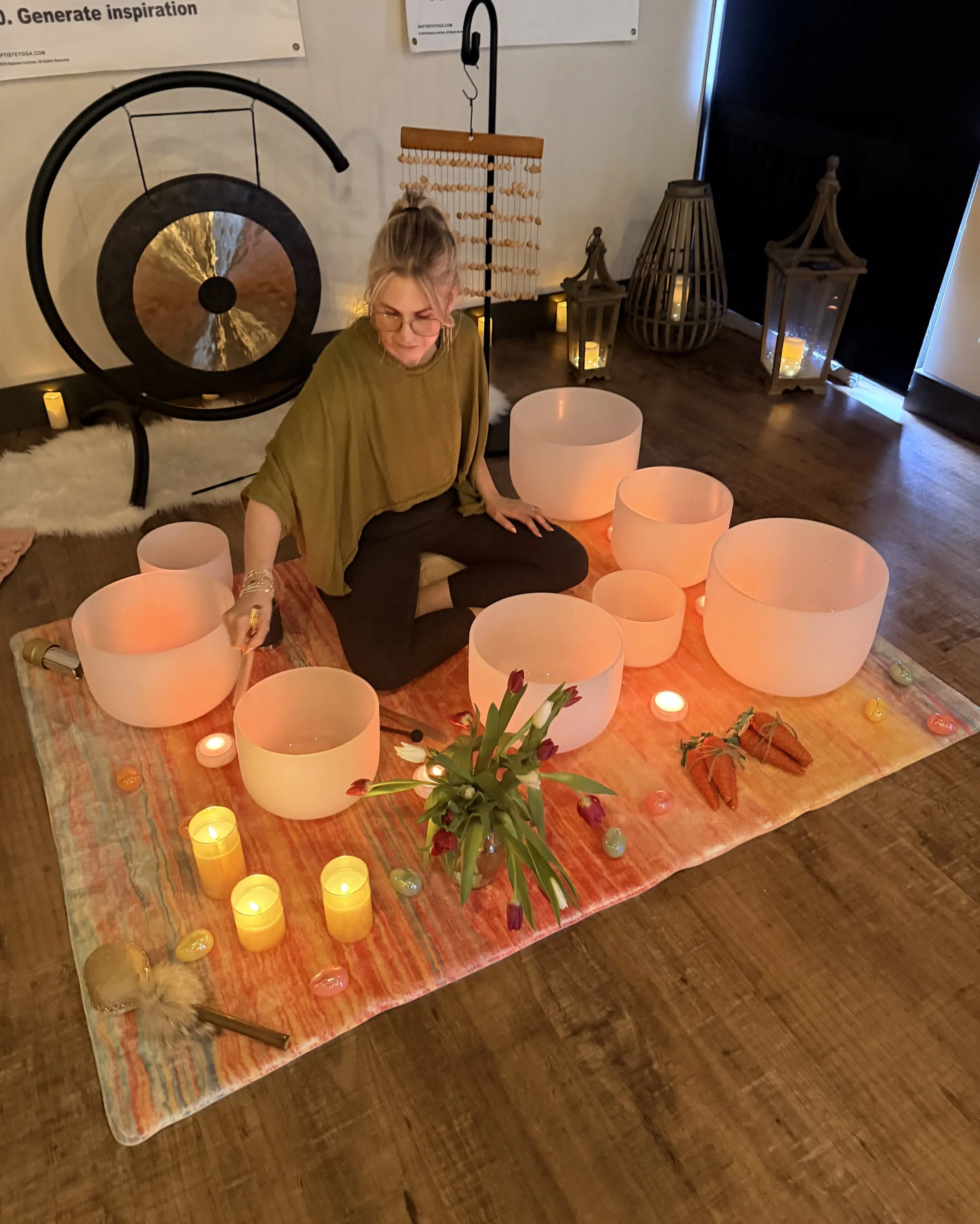 A person sitting on a colorful rug surrounded by illuminated singing bowls, candles, and flowers, with decorative lanterns and a wooden abacus in the background.