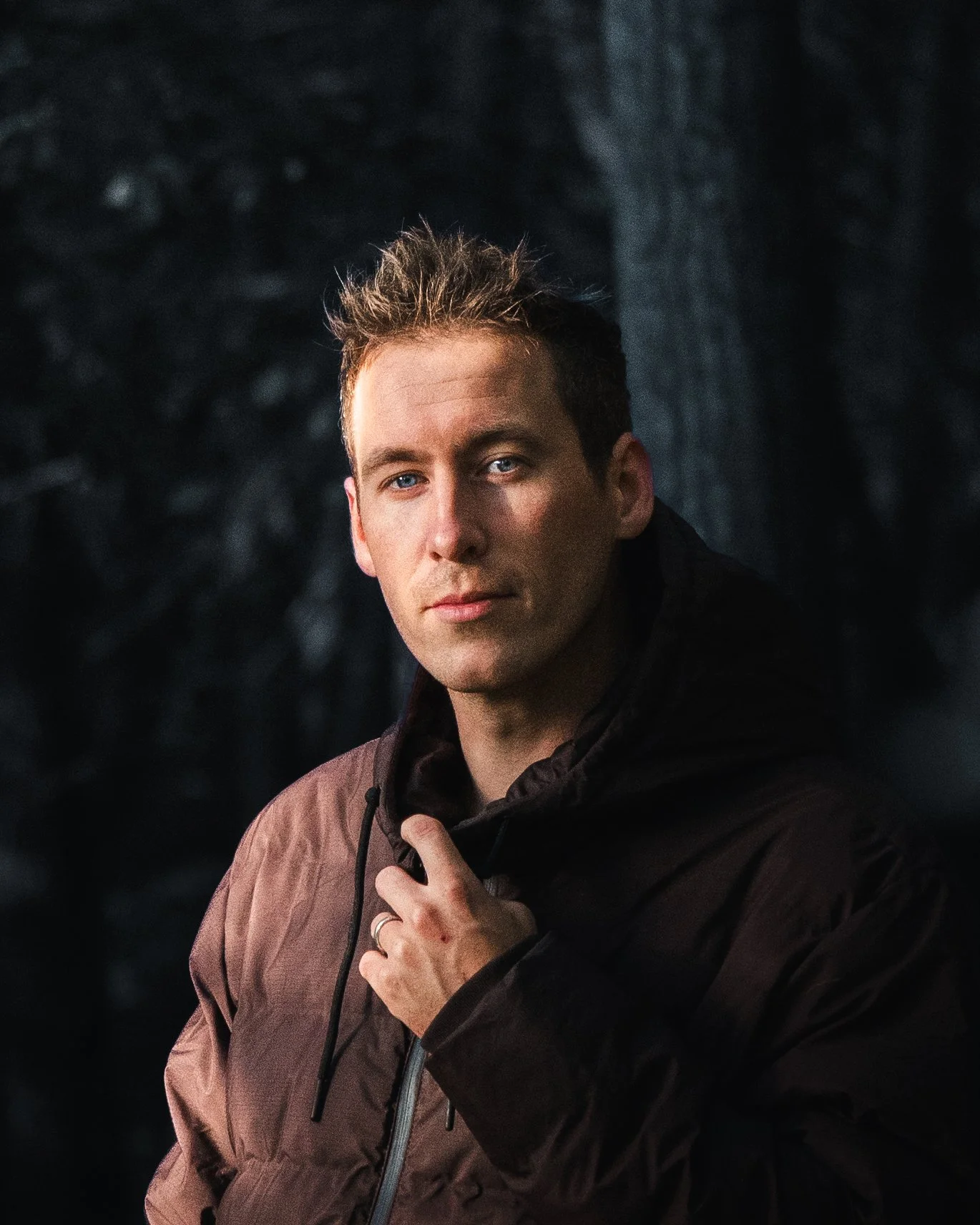 Junge Mann mit brauner Jacke und dunklem Sweatshirt vor dunklem Hintergrund.