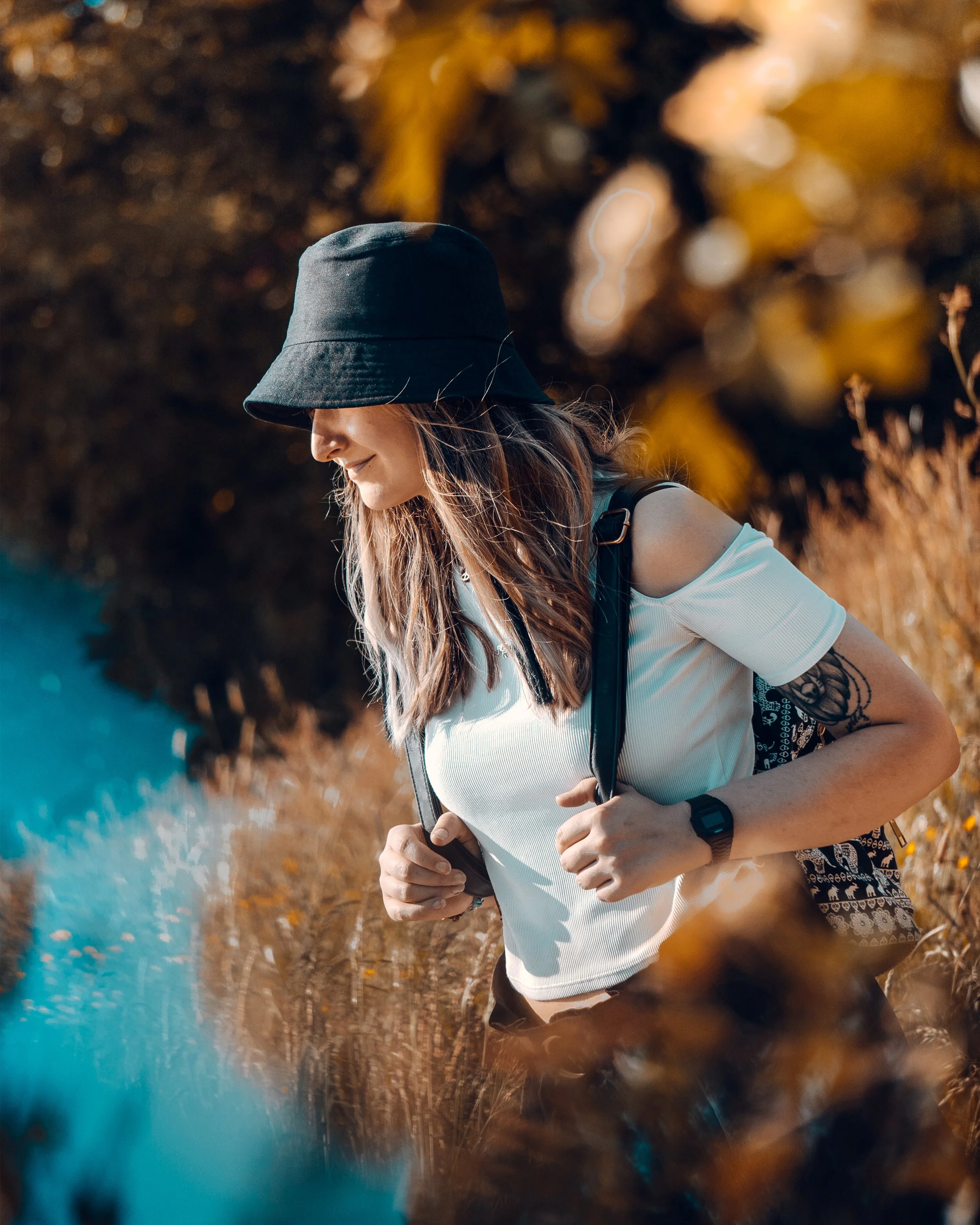 Eine junge Frau mit langem Haar trägt einen schwarzen Hut, ein weißes T-Shirt und eine schwarze Tasche und läuft im Freien durch eine herbstliche Landschaft mit bunten Blättern. Sie hält ein Smartphone in den Händen und lächelt leicht.