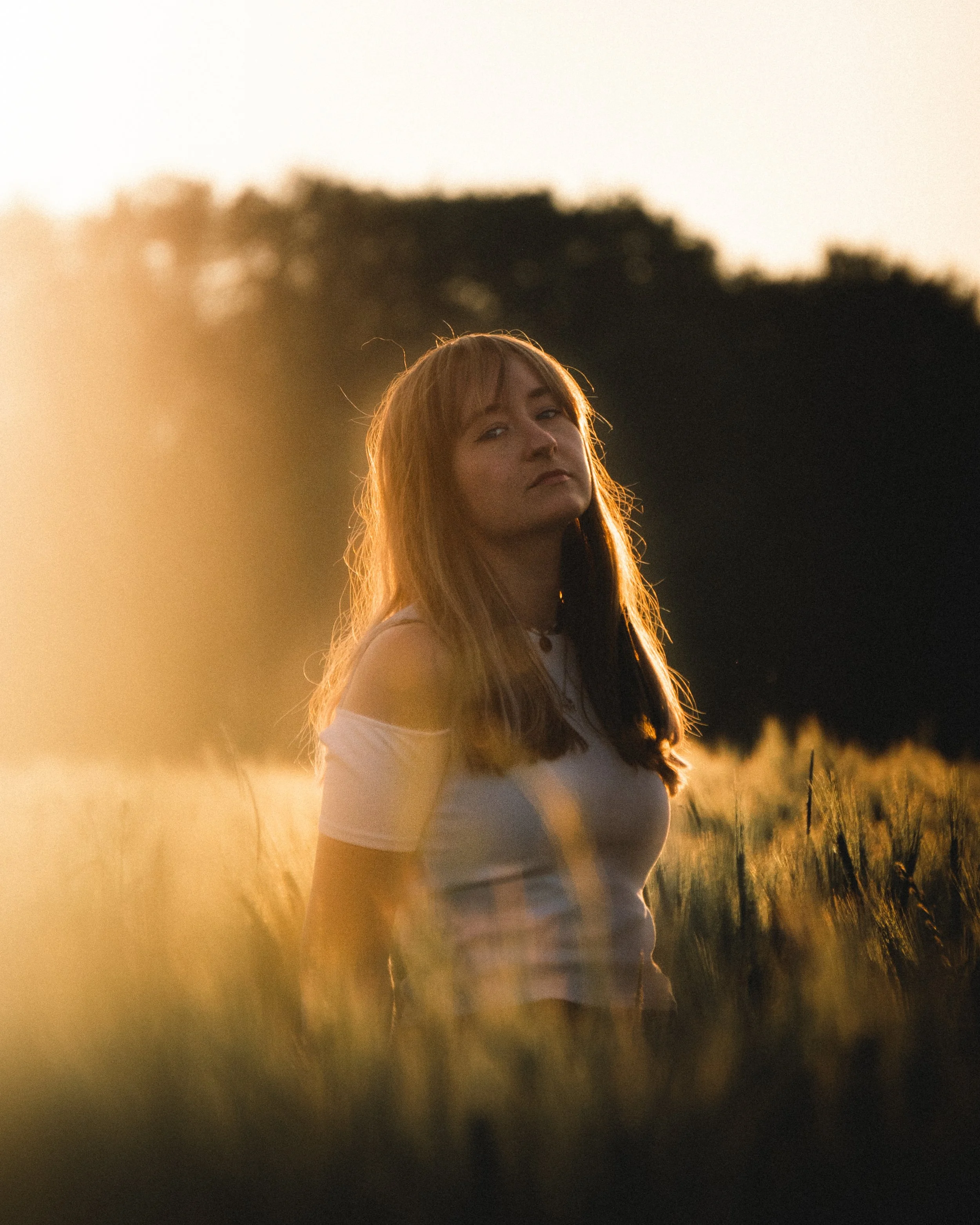 Junge Frau mit langem Haar in weißem T-Shirt steht im Sonnenuntergang auf einem Feld.