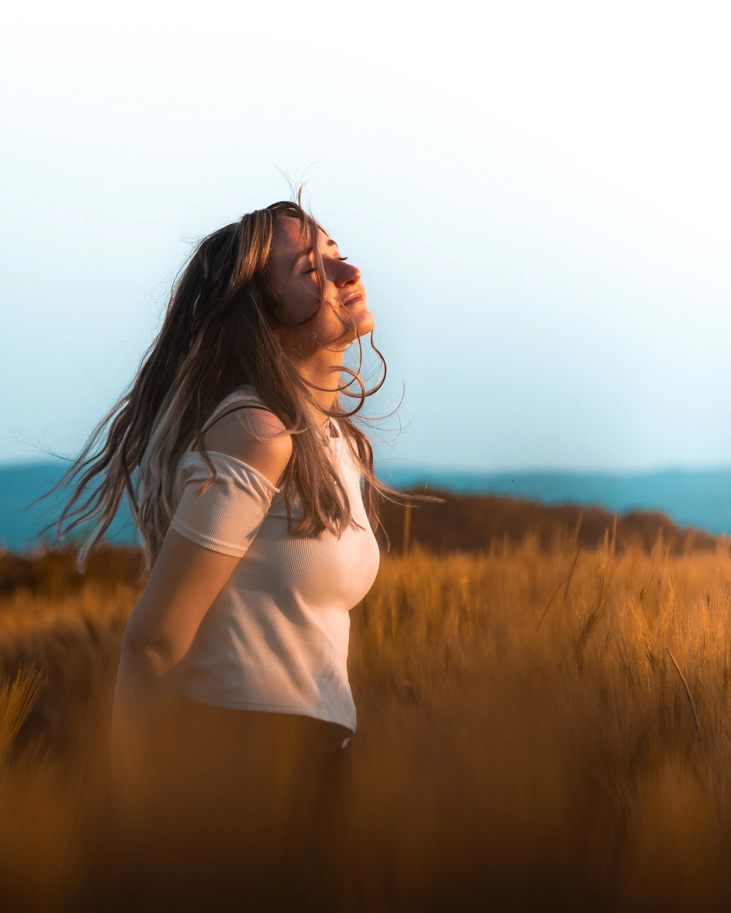 Eine junge Frau steht im Freien auf einer Lichtung während der goldenen Stunde und genießt die Sonne mit geschlossenen Augen, die Haare im Wind bewegend, umgeben von hohen Gräsern und einem klaren Himmel.