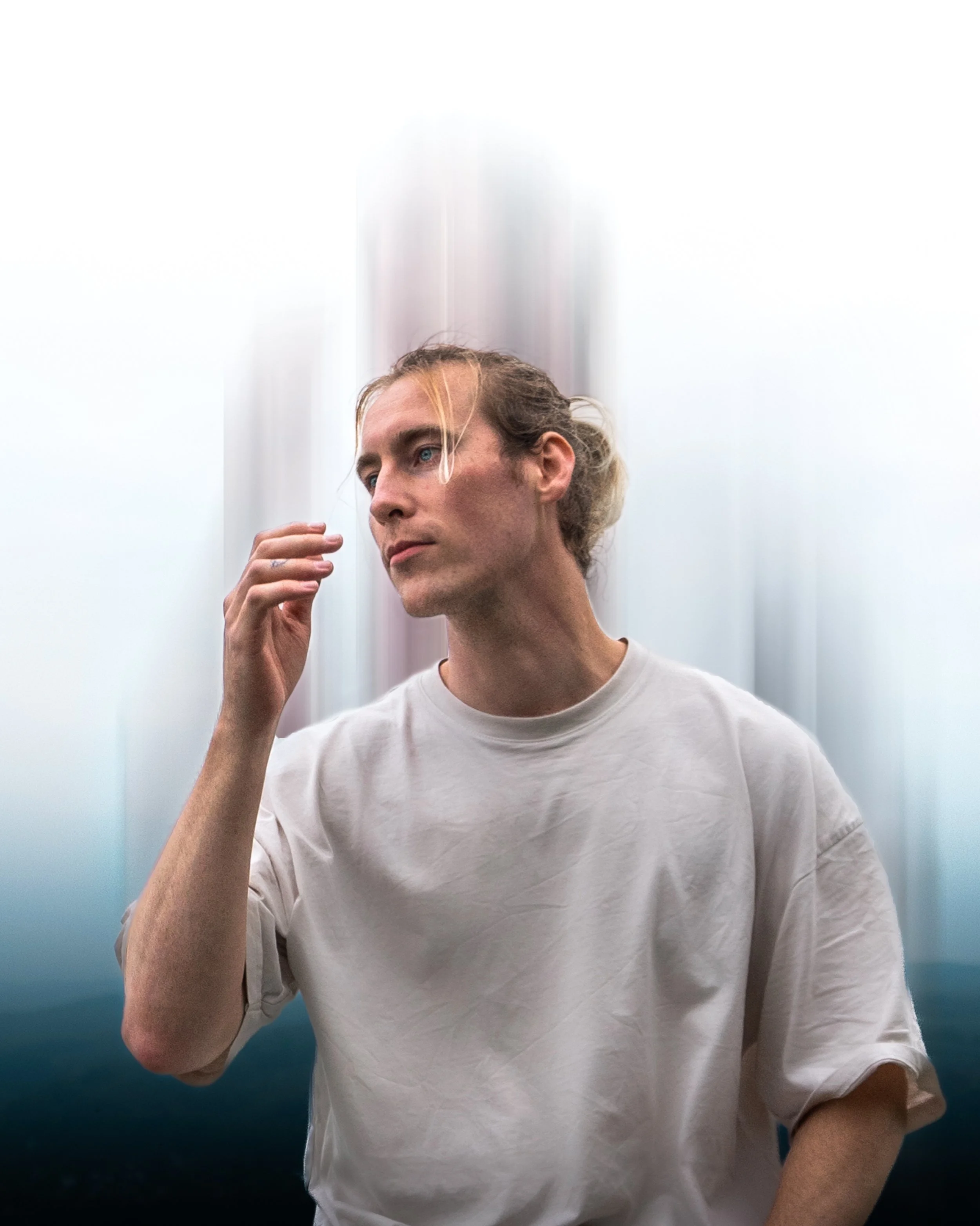 Nahaufnahme eines manns mit blonden, gewellten Haaren, der ein weißes T-Shirt trägt und leicht an die Stirn fasst. Der Hintergrund ist verschwommen und hell.
