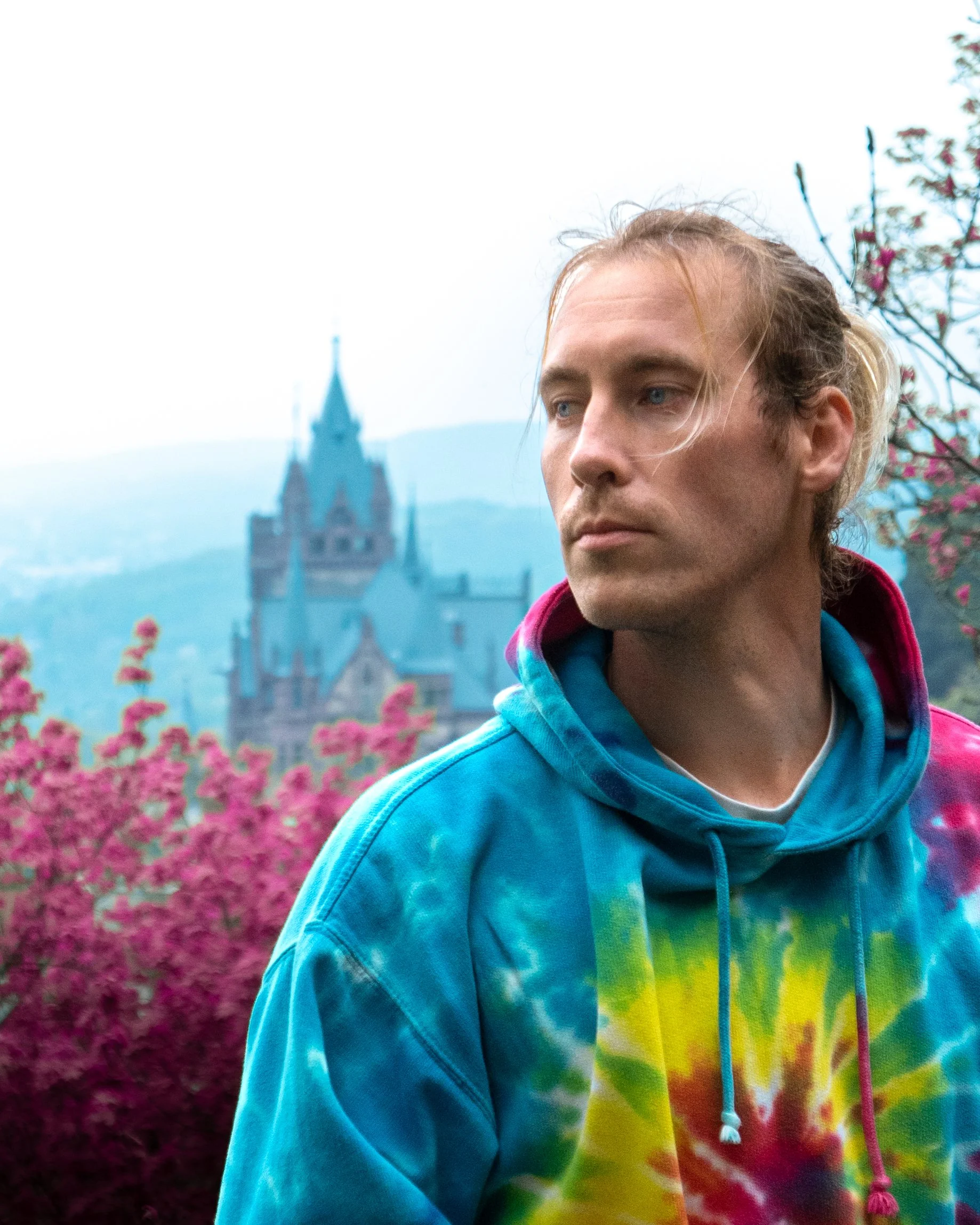 Junge Person mit blonden, lockigen Haaren, die eine bunte, farbenfrohe Kapuzenjacke trägt, steht vor einem Hintergrund mit rosa Blumen und einem Schloss im Nebel.