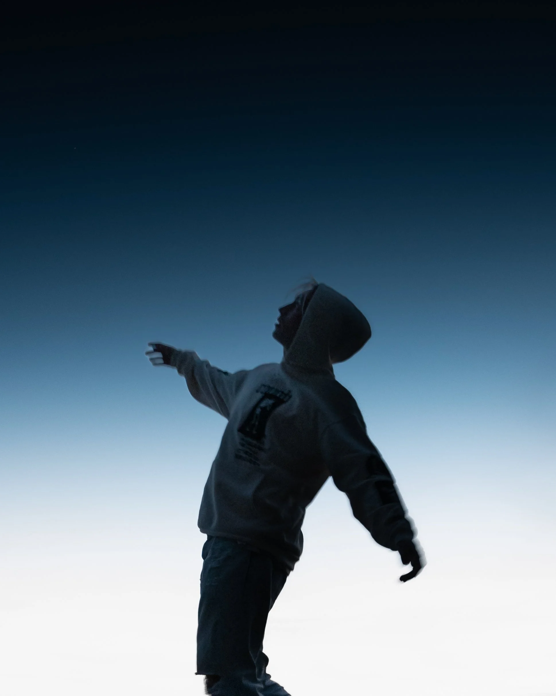 Person in einem grauen Hoodie steht vor einem klaren, blauen Himmel bei Sonnenuntergang und macht eine Geste mit der Hand, während sie nach oben blickt.