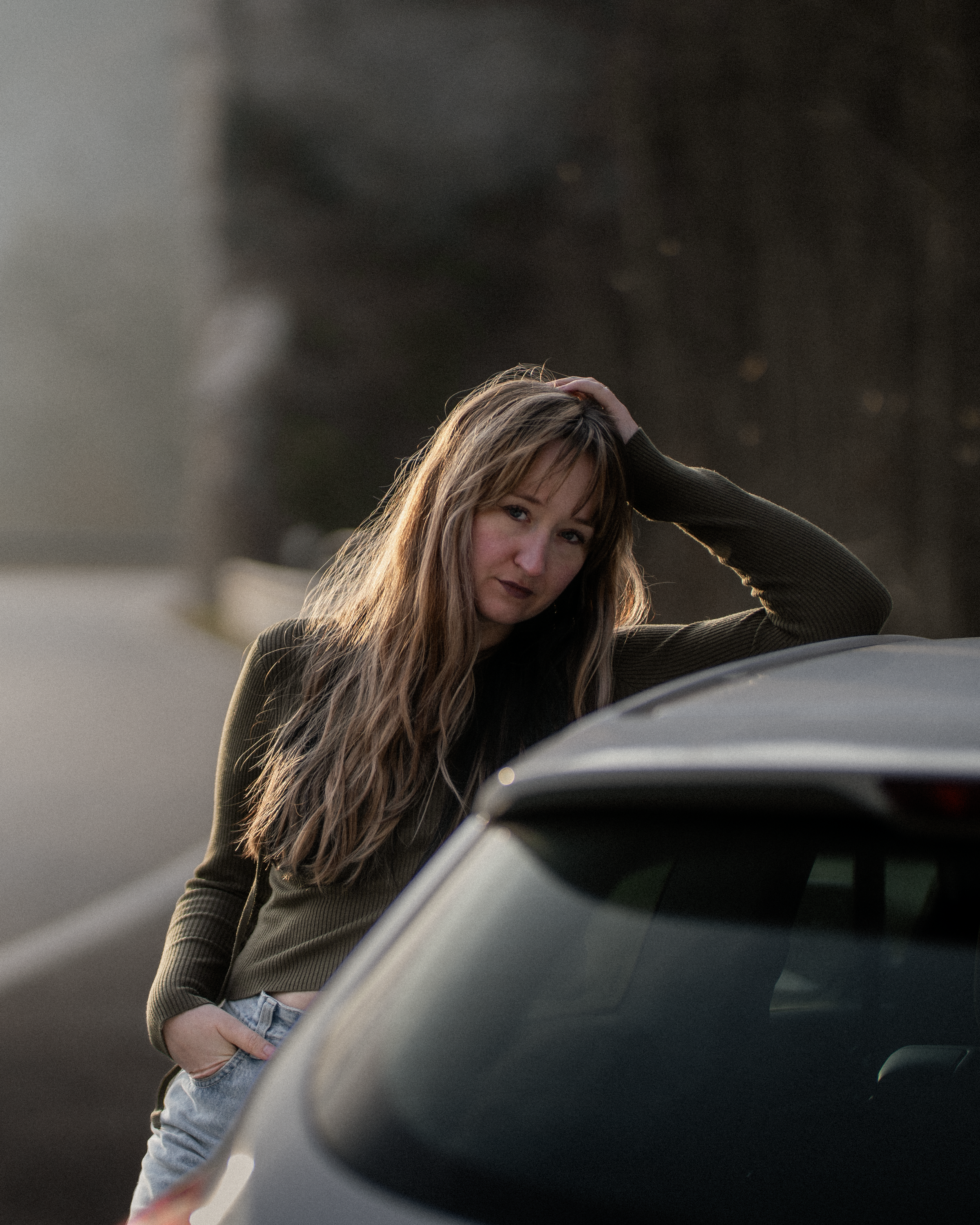 Eine junge Frau mit langen, braunen Haaren und einem dunklen Oberteil lehnt sich an ein Auto, während sie an der Motorhaube steht und in die Kamera schaut. Die Umgebung ist unscharf, vermutlich ein Außenbereich bei Sonnenuntergang.