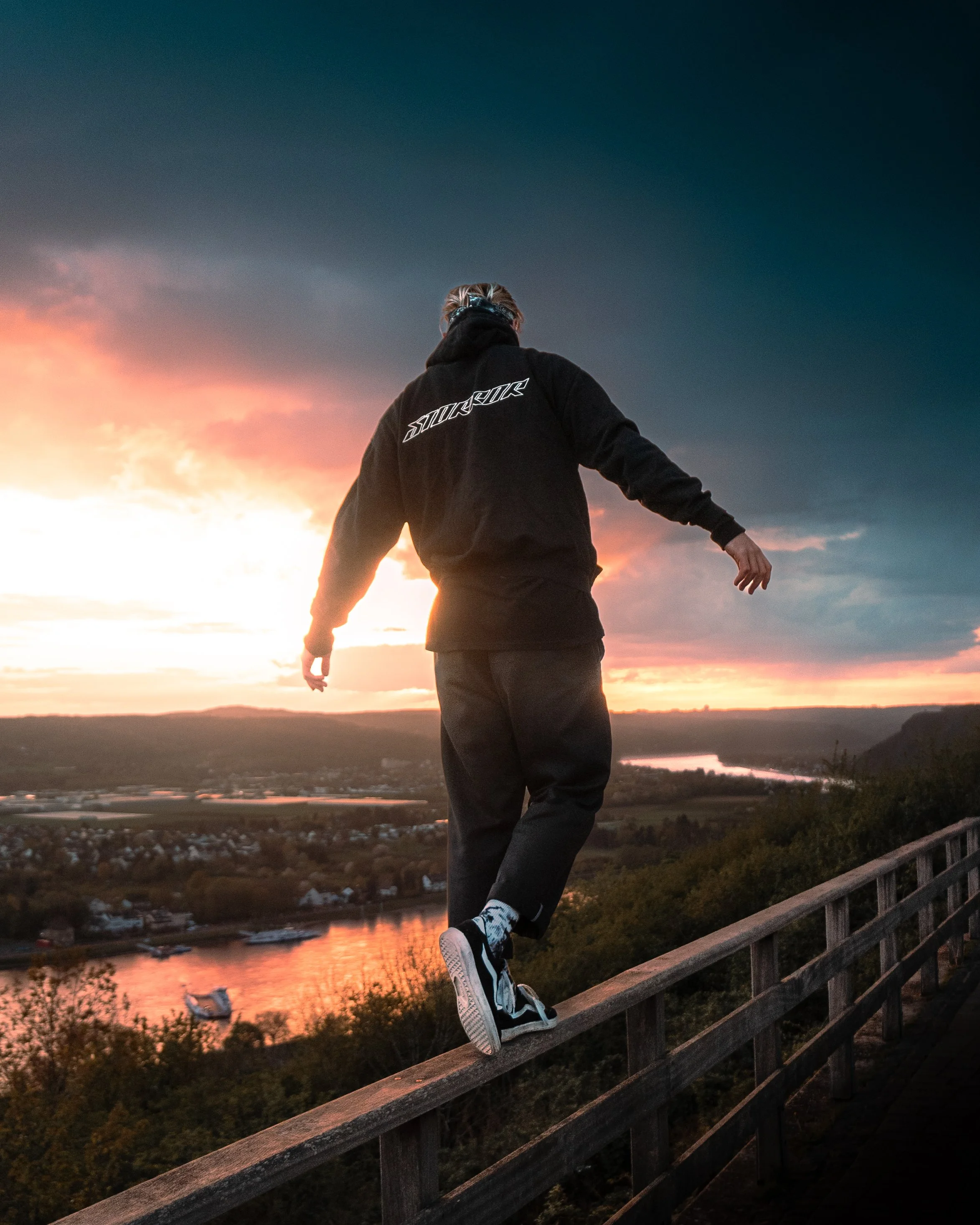 Junger Mann sitzt auf einem Holzgeländer an einem Fluss, während die Sonne untergeht und der Himmel in Orange und Blau leuchtet.