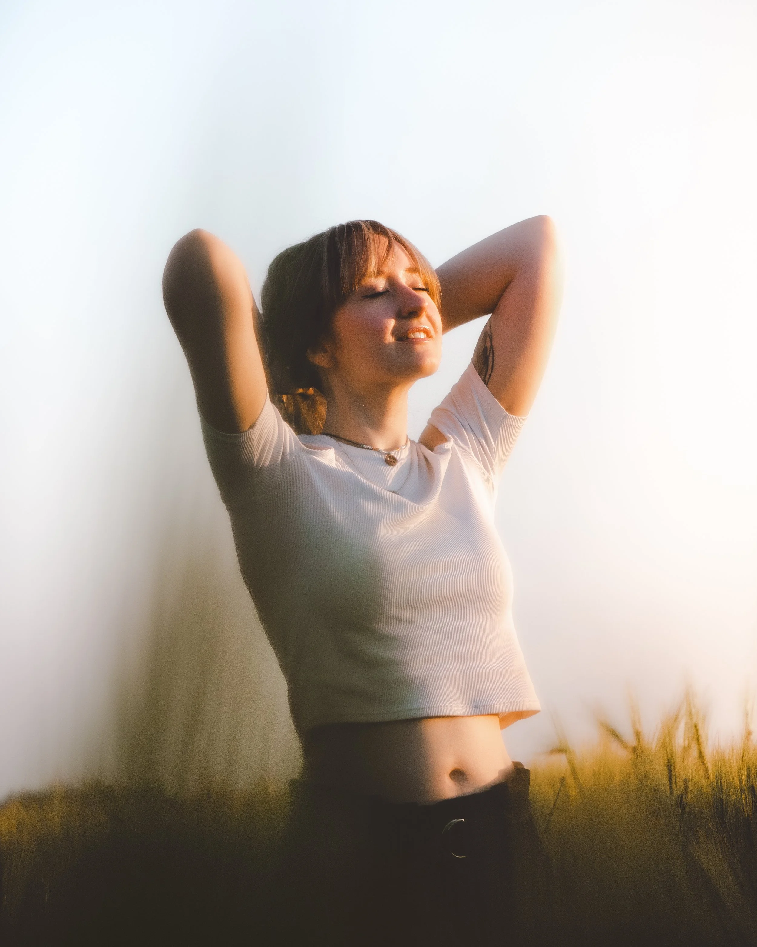 Junge Frau mit Kurzhaarfrisur, die draußen bei Sonnenuntergang mit den Augen geschlossen und einem Lächeln im Gesicht steht, mit den Armen hinter dem Kopf.