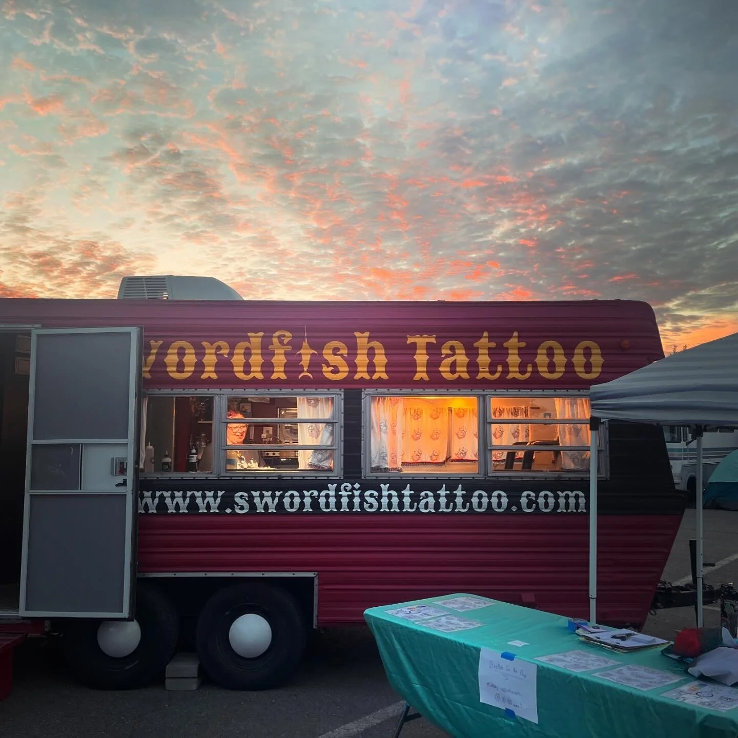 A food truck named 'swordfish Tattoo' parked outdoors during sunset, with a table in front displaying flyers and materials.