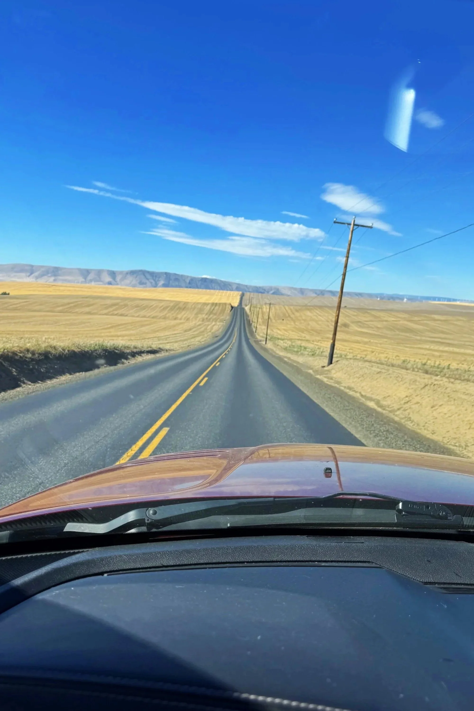 Photo taken from inside a vehicle looking out through the windshield at a long, straight desert road with yellow fields on both sides, utility poles lining the road, and distant mountains under a clear blue sky with scattered clouds.