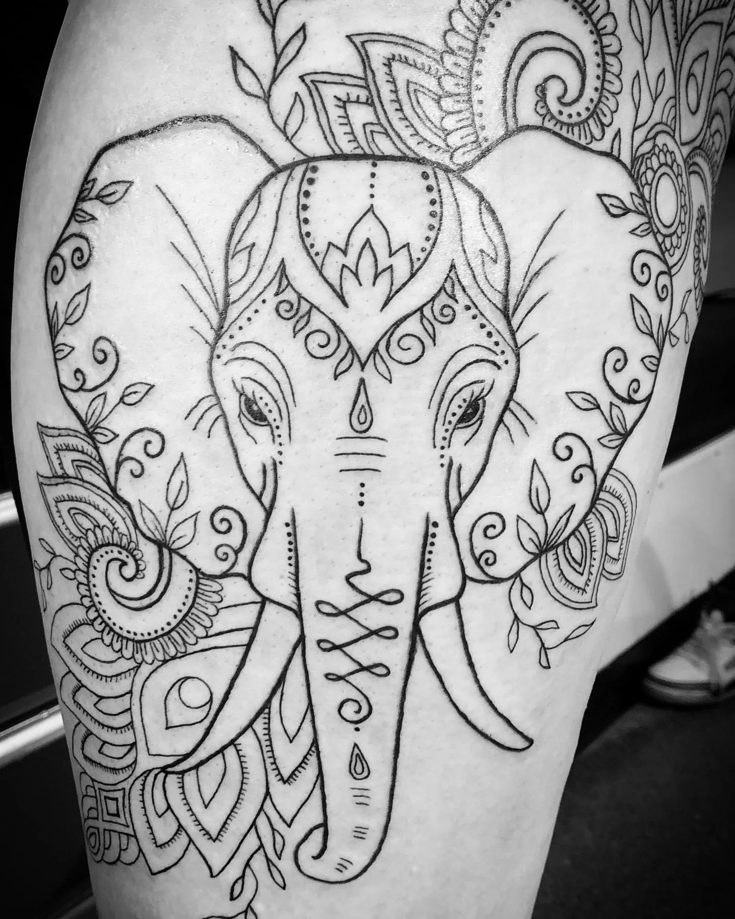 Black and white tattoo outline of an elephant surrounded by decorative patterns and floral elements.